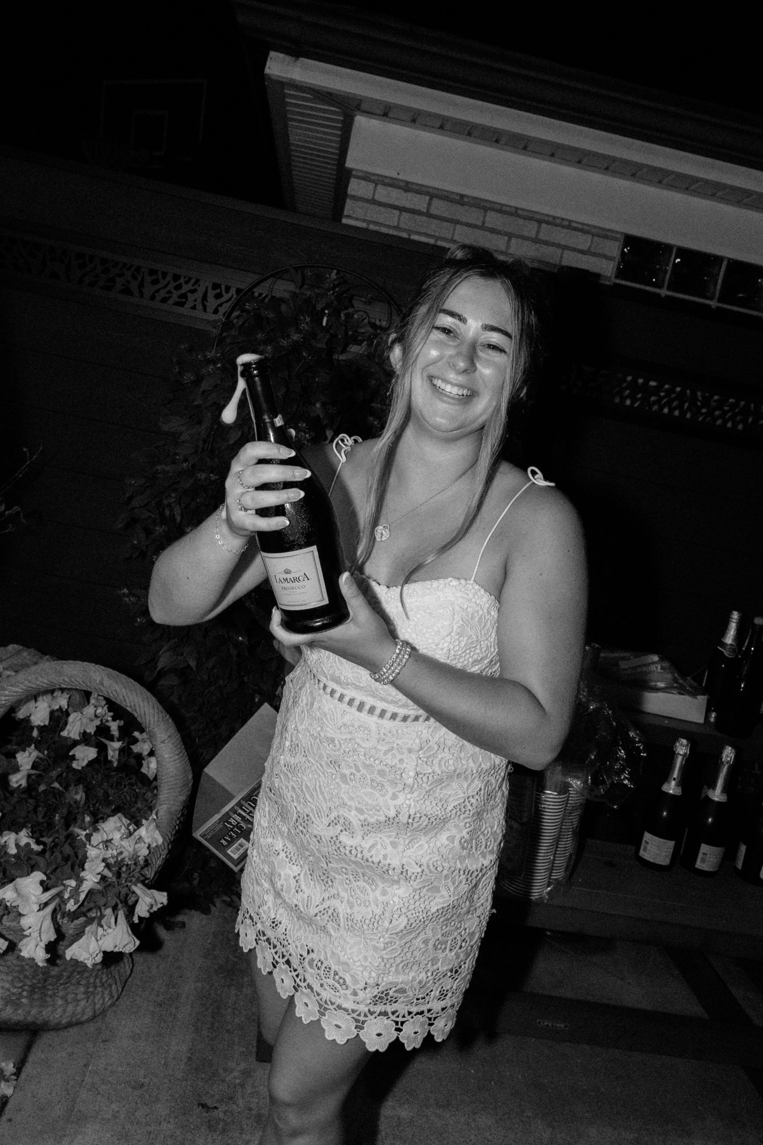 Woman in white lace dress smiling and holding a wine bottle at an outdoor evening party.
