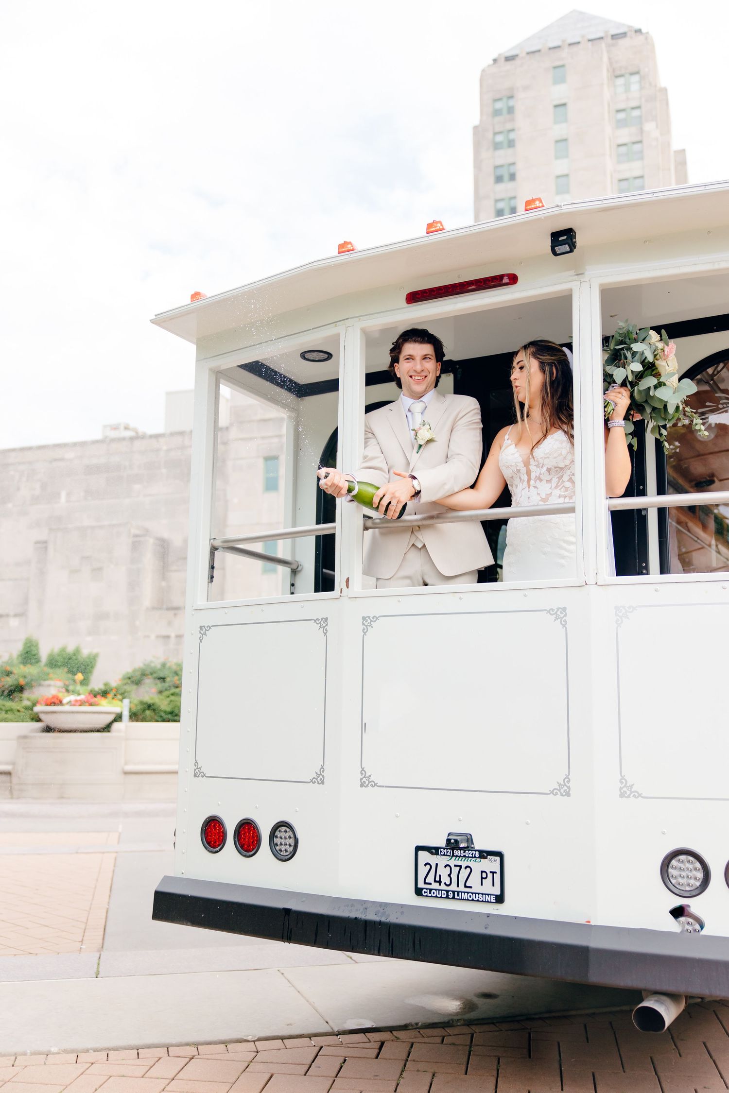 Bride and groom celebrate on a white trolley car, popping champagne in front of a city building.