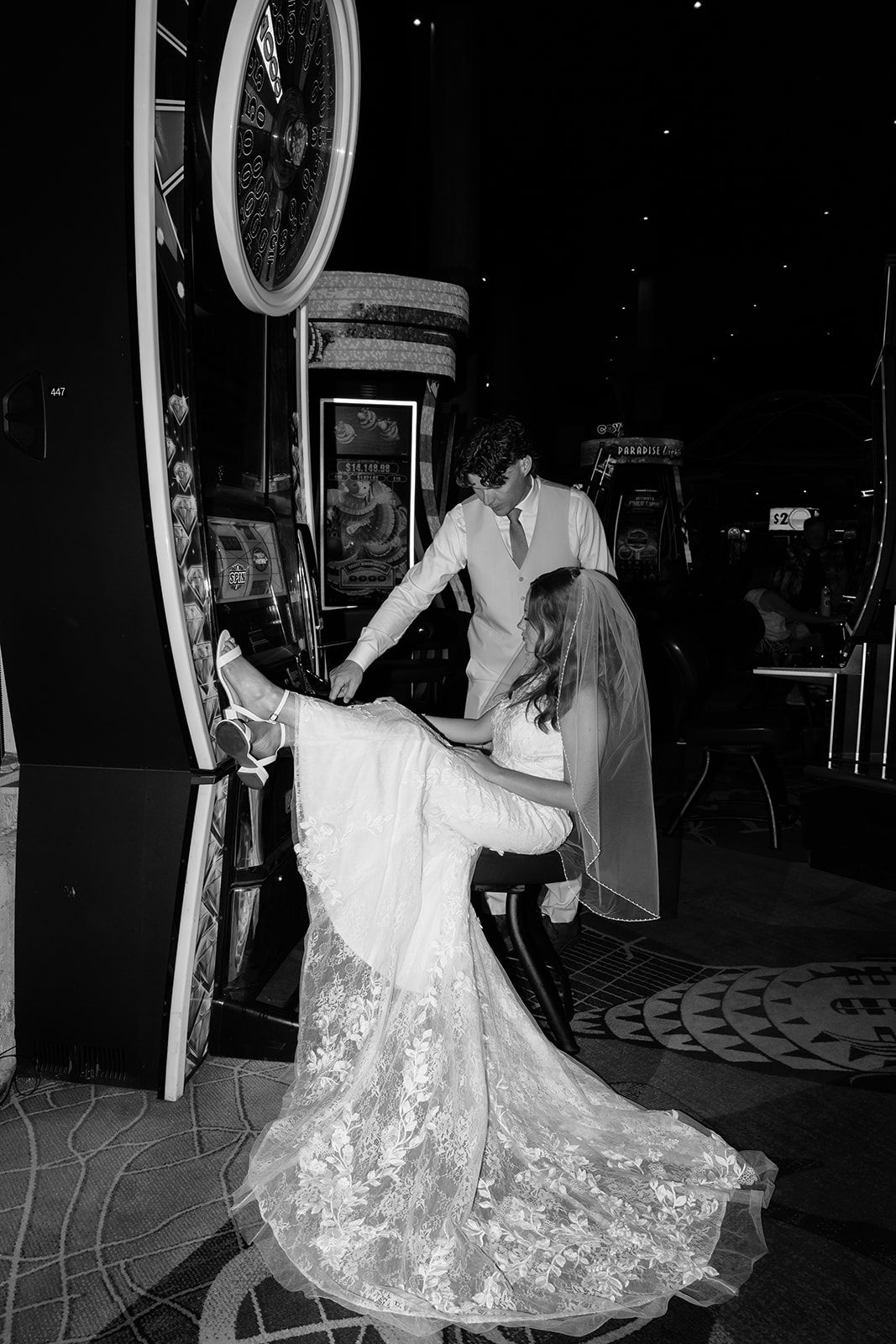 Bride and groom play slot machines in casino on their wedding day, bride wearing lace gown and veil.