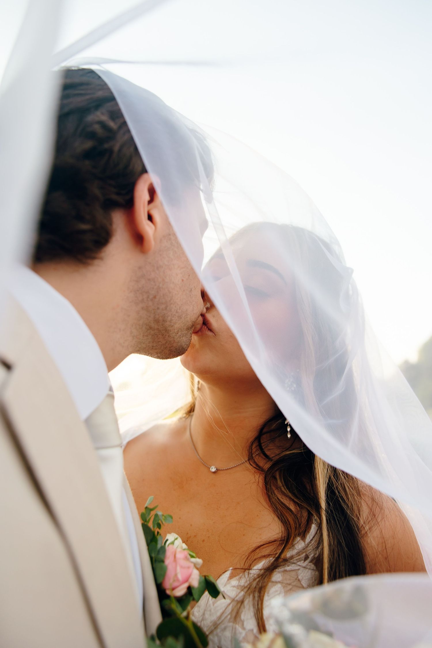 Bride and groom sharing a romantic kiss under a flowing white veil, bride holding pink roses bouquet.
