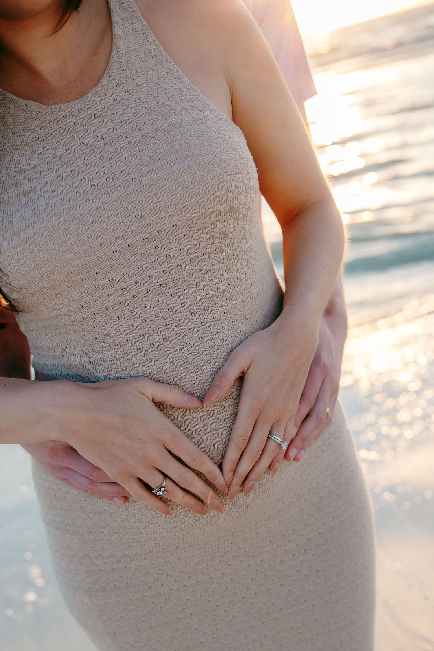 Pregnant woman in beige dress with couple forming heart shape with hands on baby bump at sunset beach.