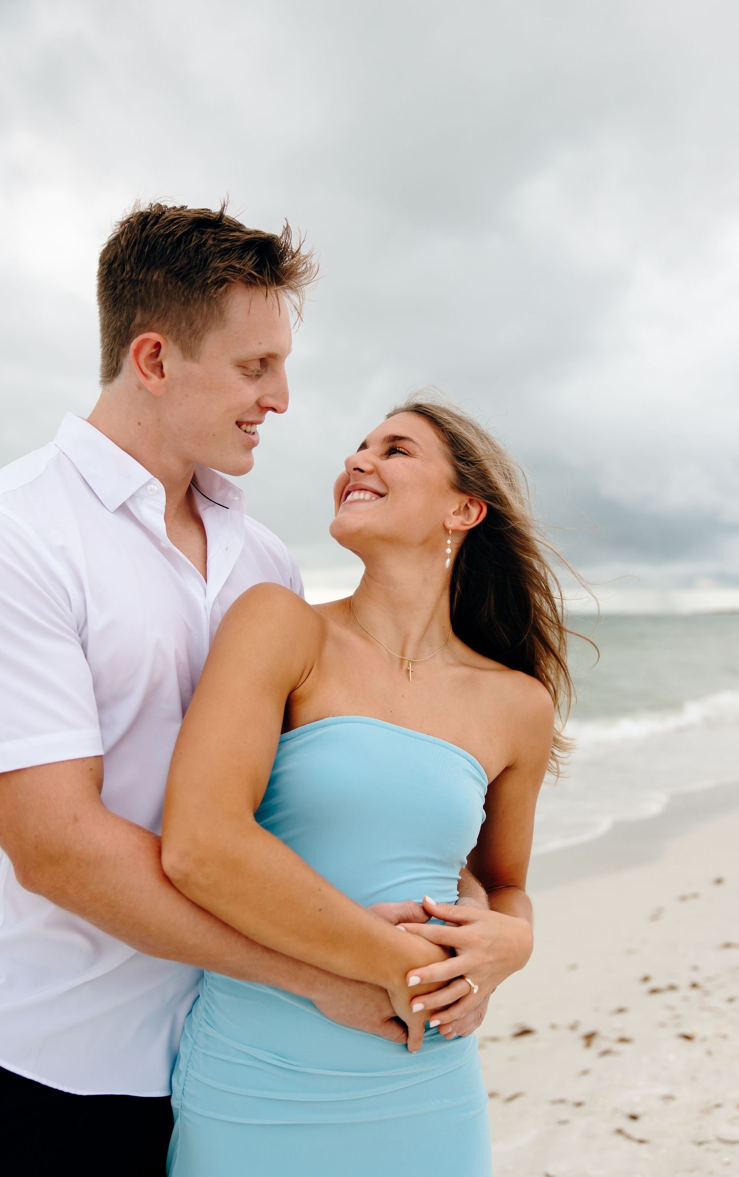 A romantic couple embraces and laughs together on a cloudy beach while wearing white and turquoise clothing.