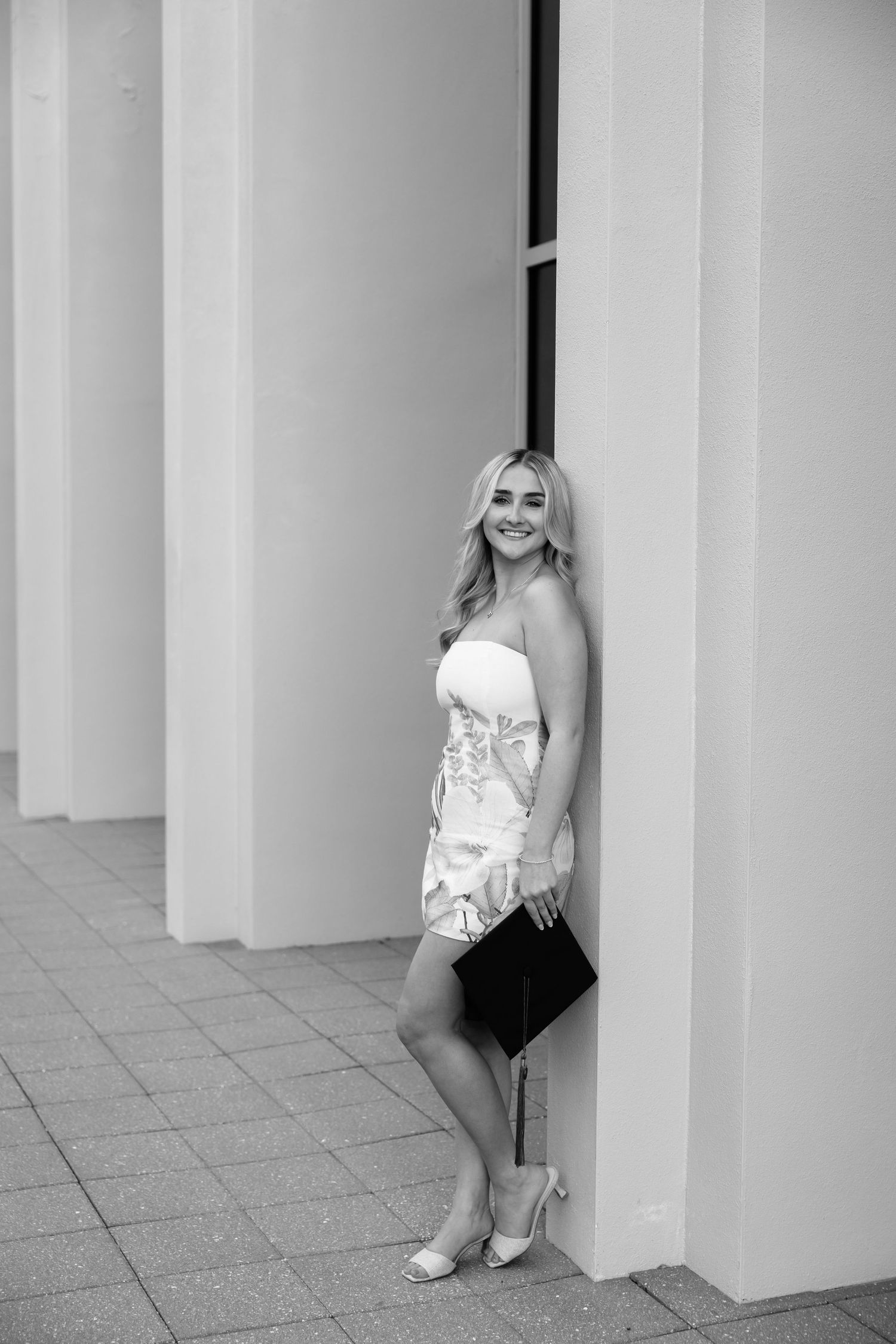 Young blonde woman in strapless dress holding clutch purse, posing elegantly between white columns in black and white photo.