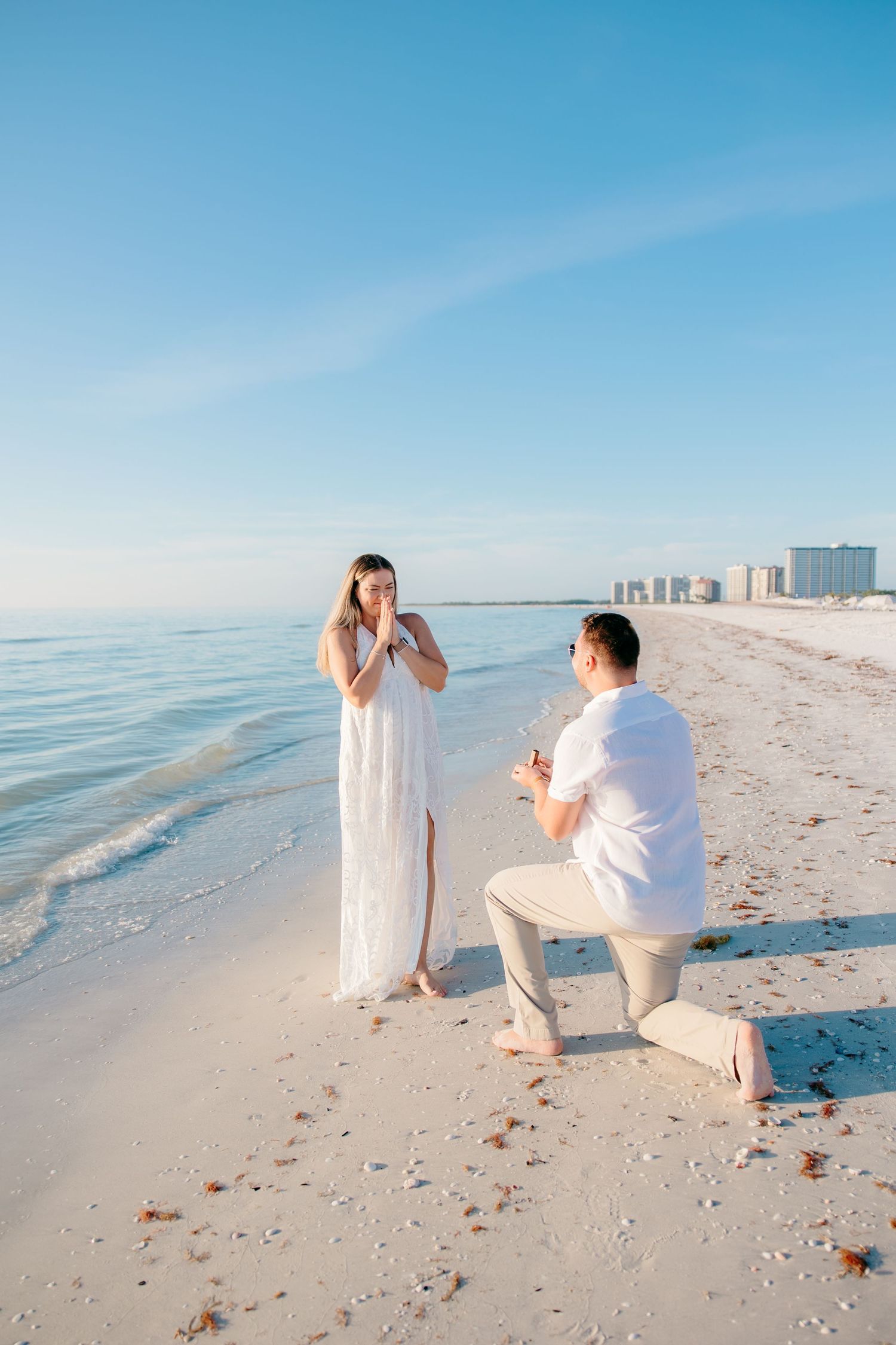 A romantic beach proposal scene unfolds in a series of photos showing a couple on white sand with blue ocean backdrop.