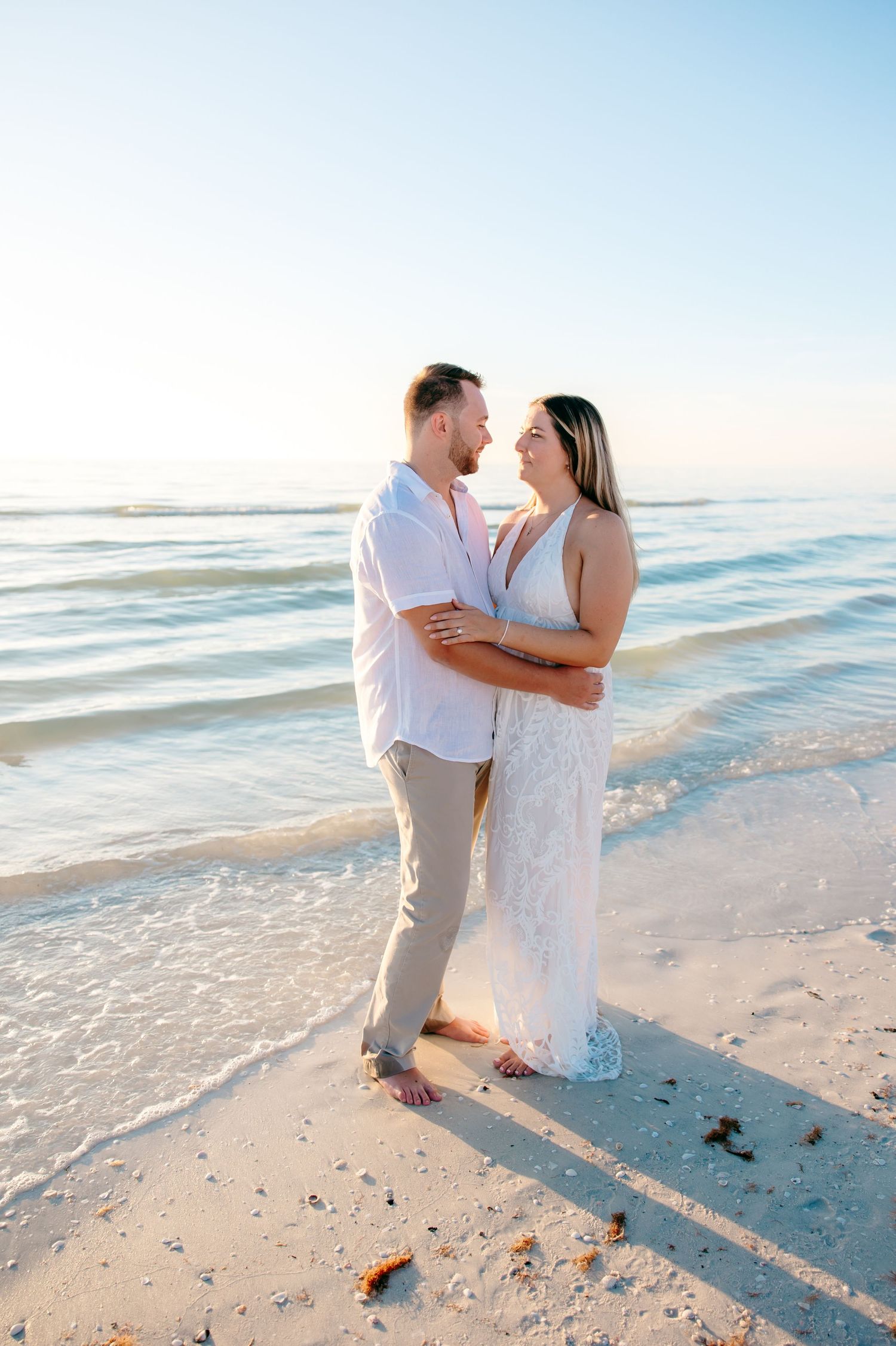 A couple embraces on a sunny beach at sunset, wearing white and beige outfits while standing in the sand near the ocean.