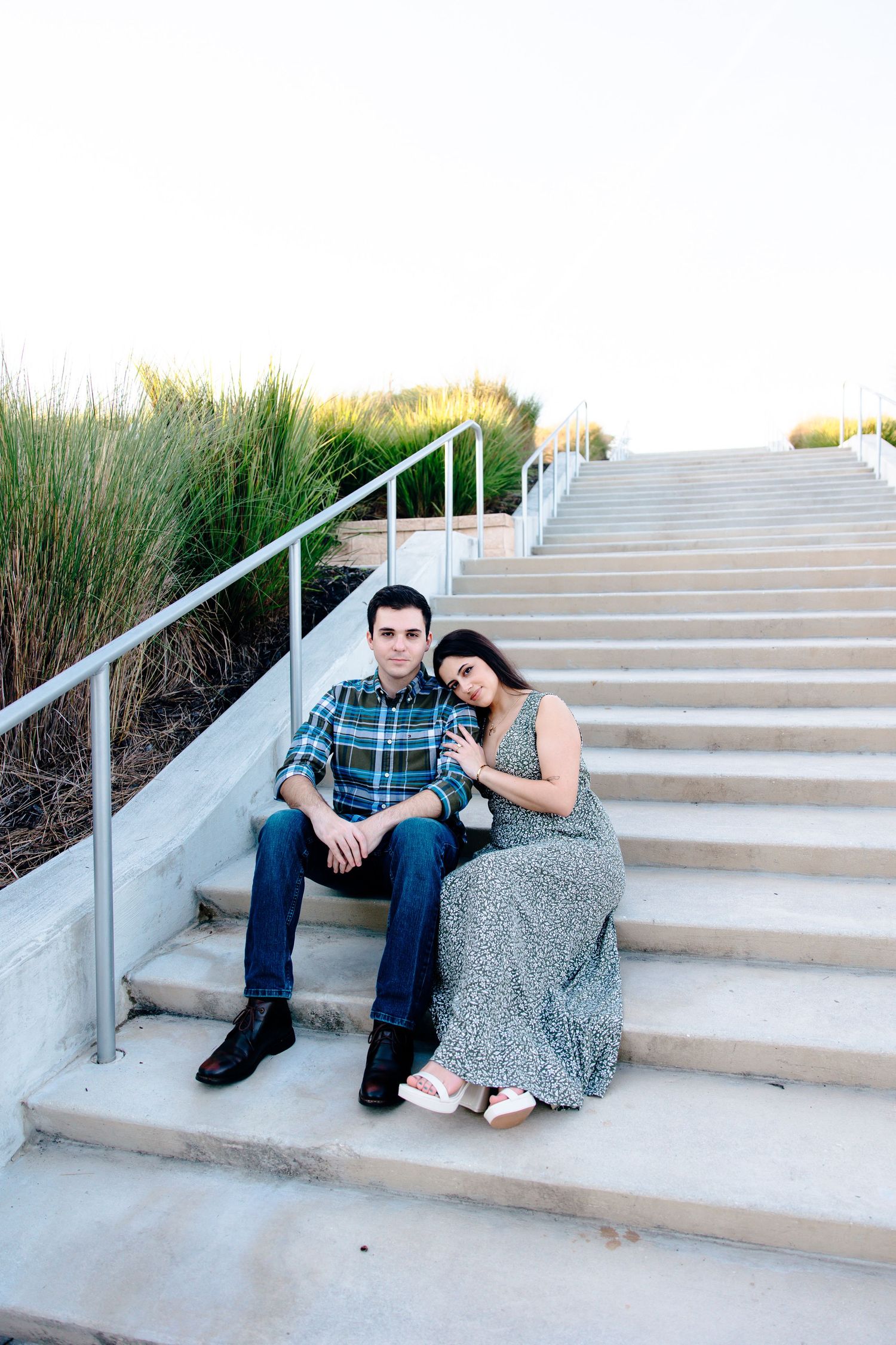 Couple sitting on outdoor concrete steps, woman in floral dress leaning on man in plaid shirt, with tall grass behind them.