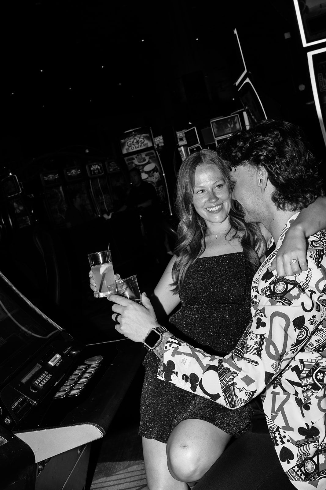 Black and white photo of smiling couple embracing at arcade bar, man in card suit kissing womans cheek.