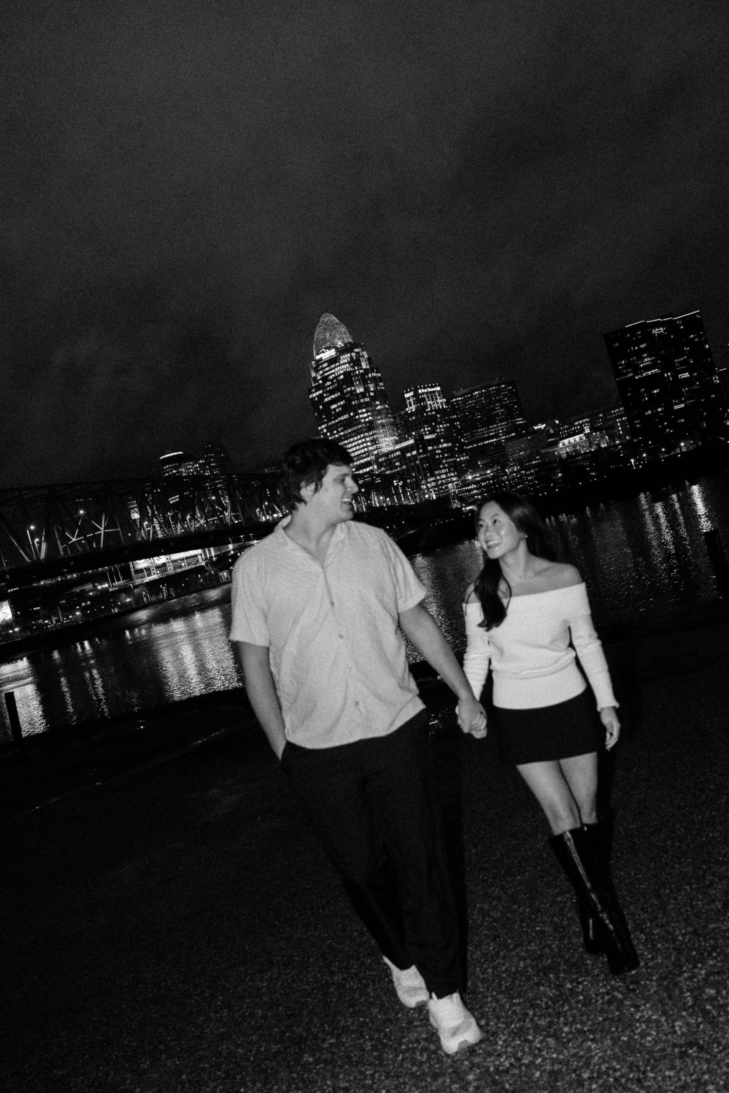 Couple holding hands walking along waterfront at night with city skyline lights reflecting on river in black and white.