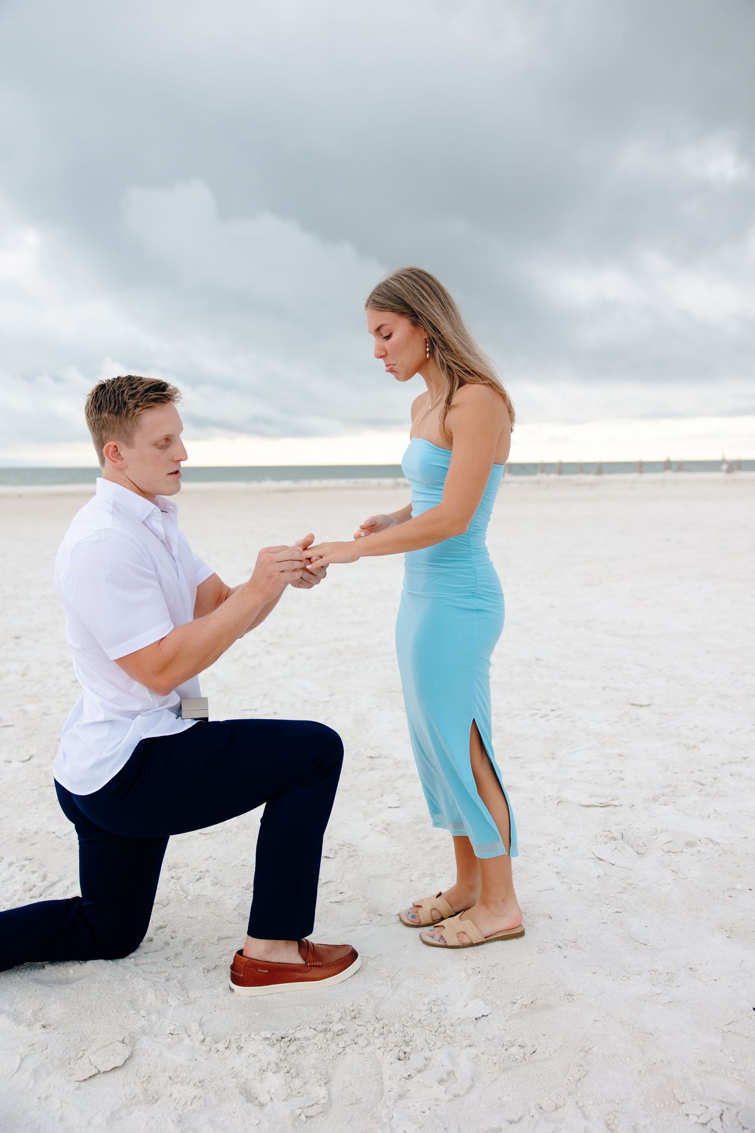 Man kneeling on one knee proposing to woman in blue dress on a sandy beach under cloudy skies.