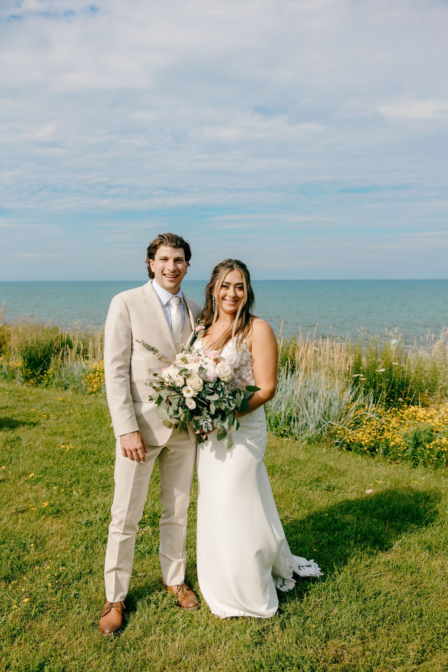 Bride and groom pose on green lawn near ocean, she holds white floral bouquet, he wears beige suit.