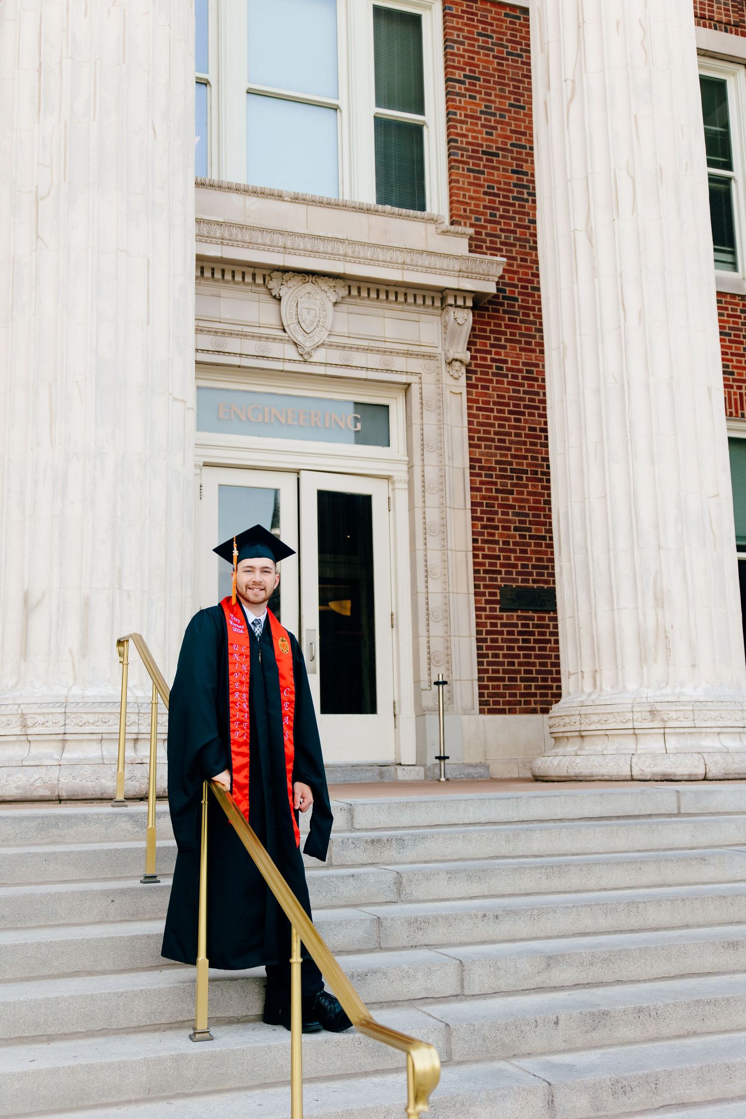 Graduate in black cap and gown with red sash poses on steps of brick and column university building.