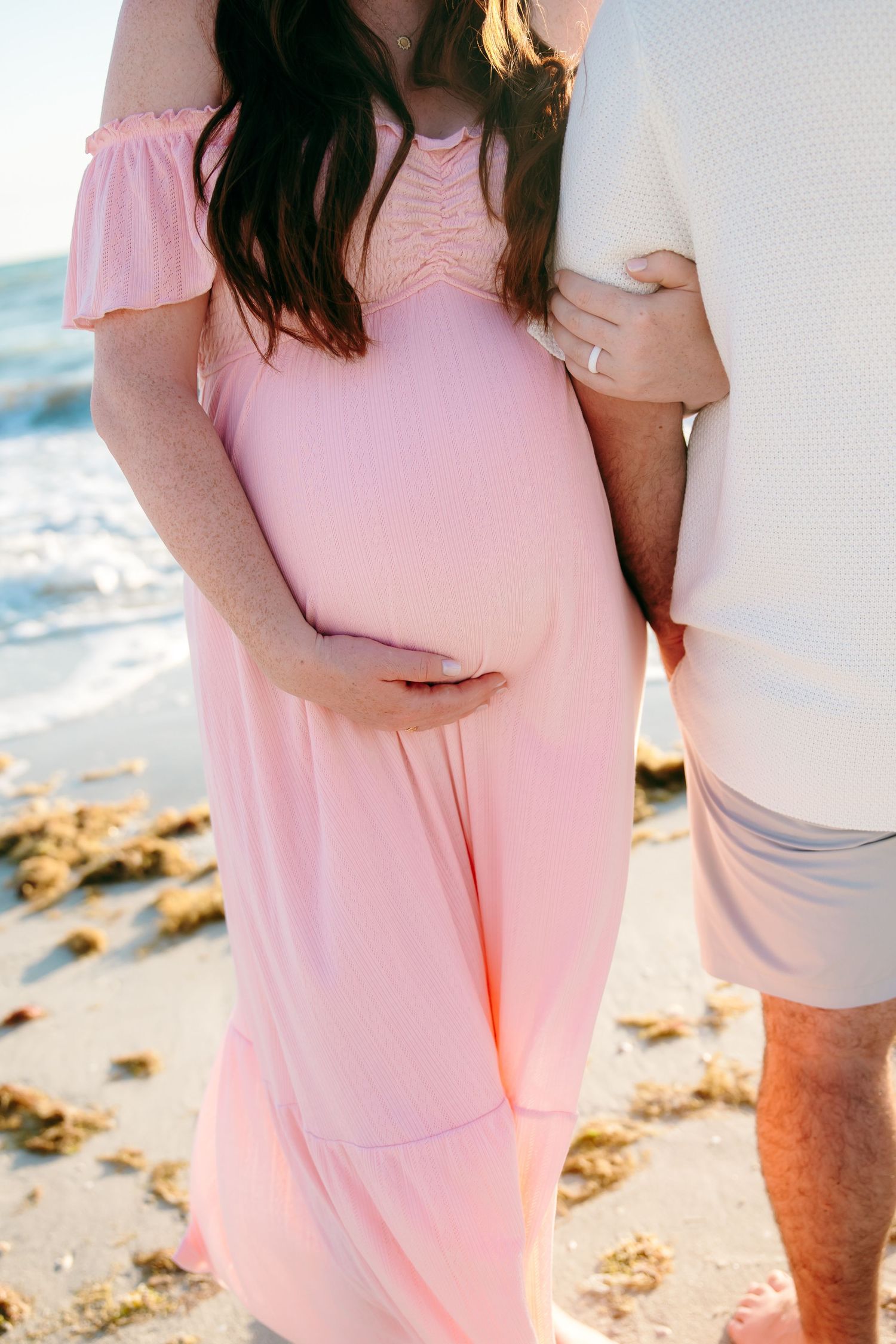 Pregnant woman in pink off-shoulder maxi dress holding baby bump on beach with partner.