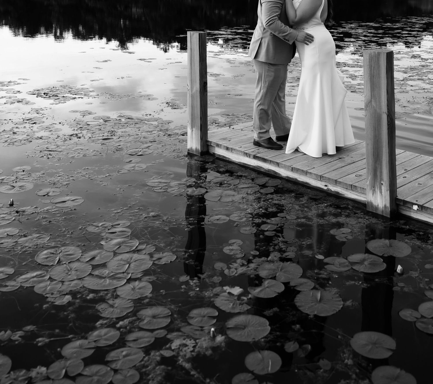 Black and white photo of a bride and groom embracing on a dock surrounded by lily pads on a calm reflective lake.