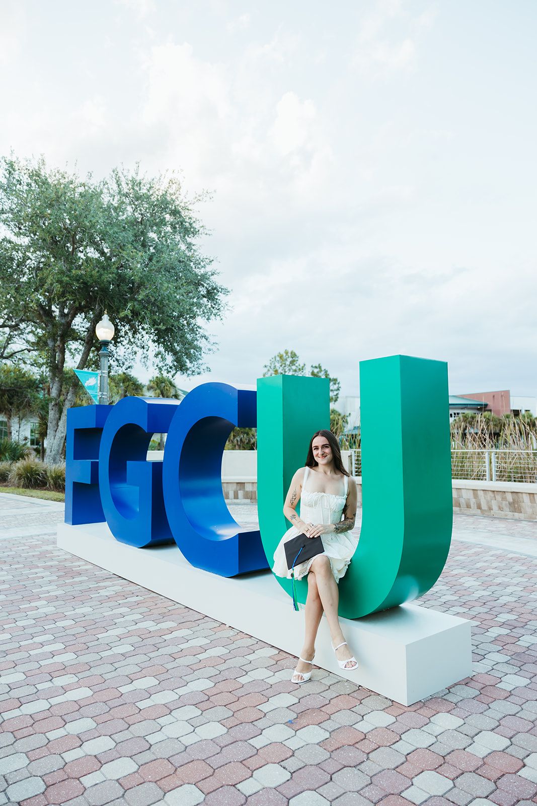 Large blue and green sculptural letters create an artistic installation on a brick paved plaza with trees in the background.