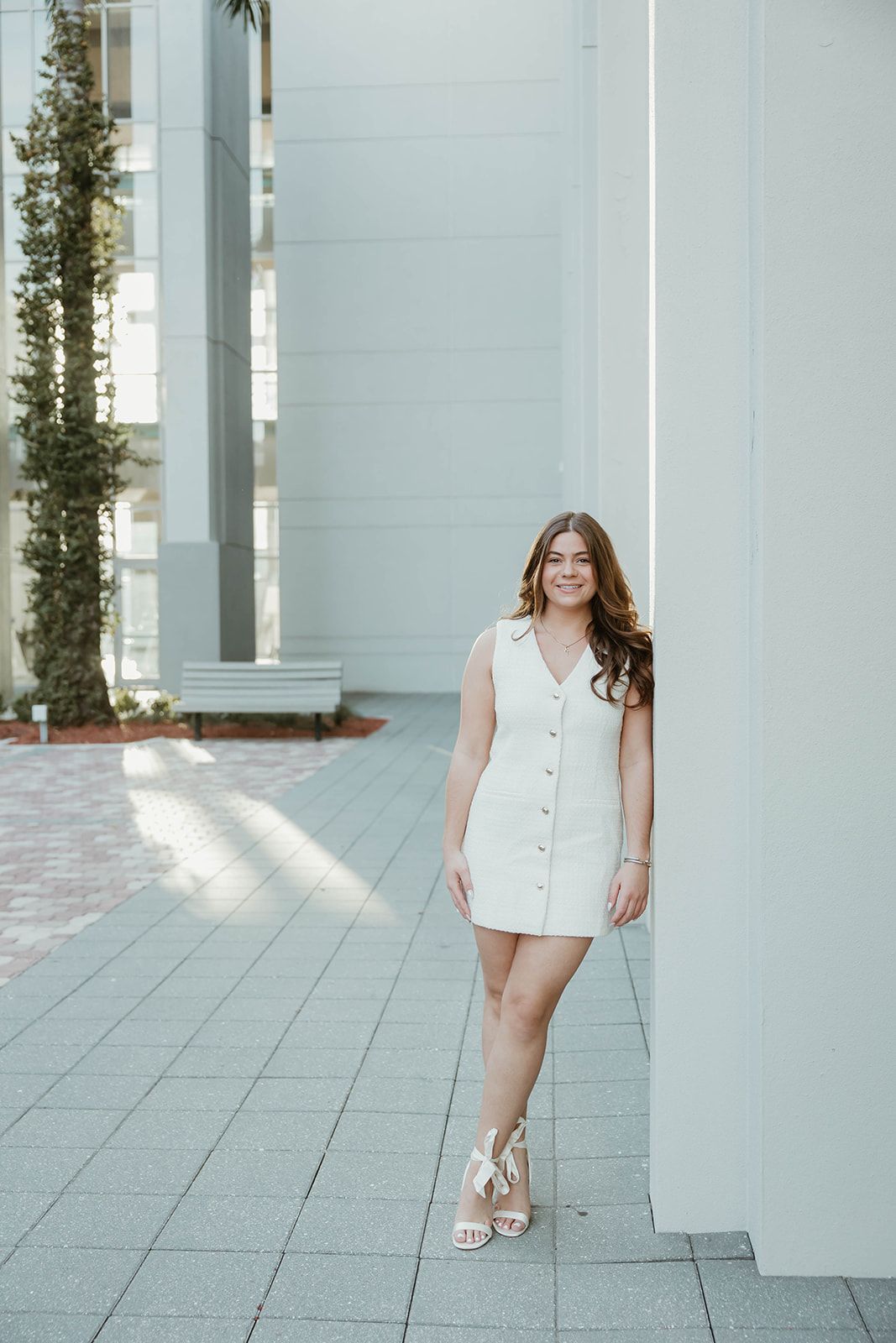 A person in a white sleeveless dress poses against a modern architectural backdrop with columns and tiled flooring.