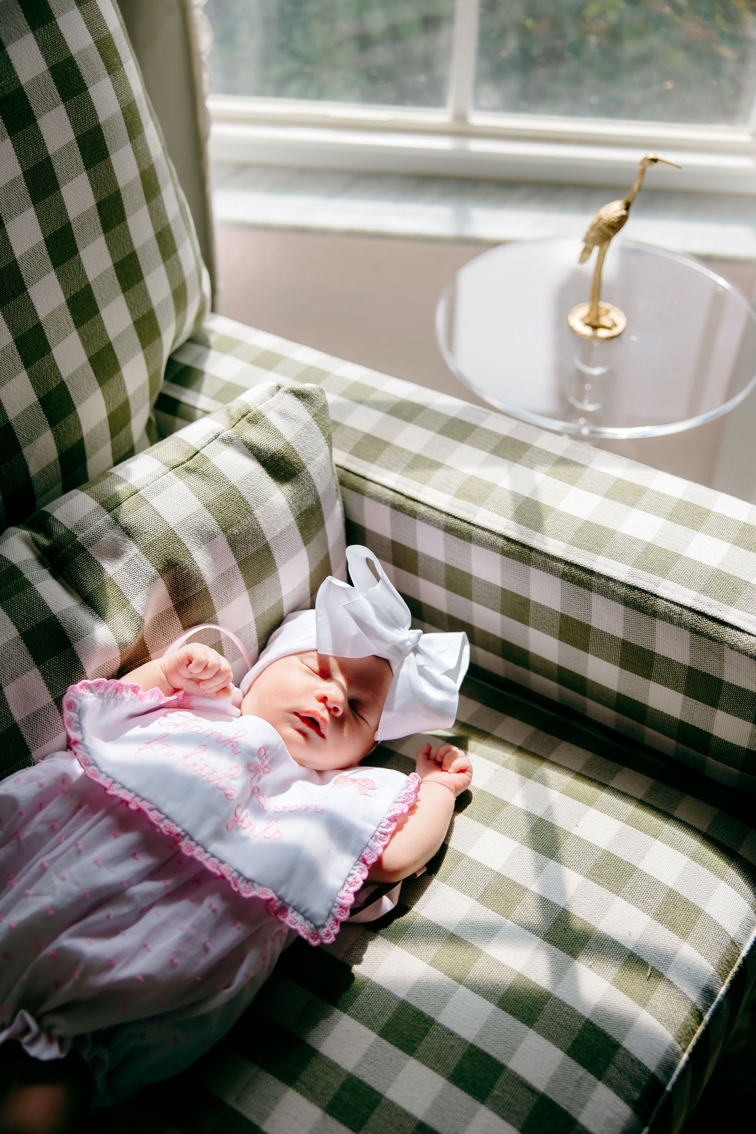 Newborn baby sleeping peacefully in a green gingham armchair wearing a pink outfit and white bow headband.