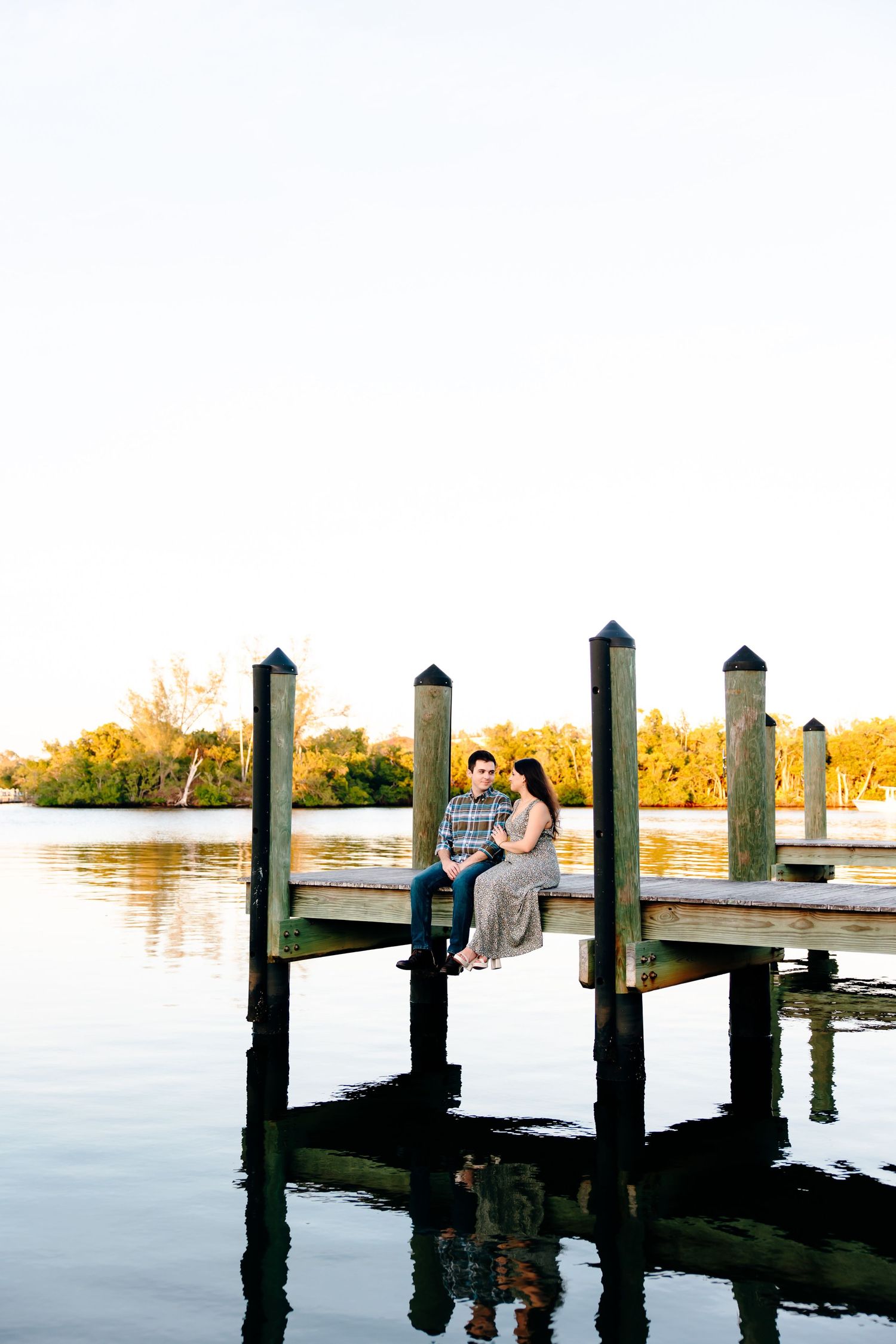 Couple sitting together on a wooden dock over calm water at golden hour, surrounded by lush greenery.