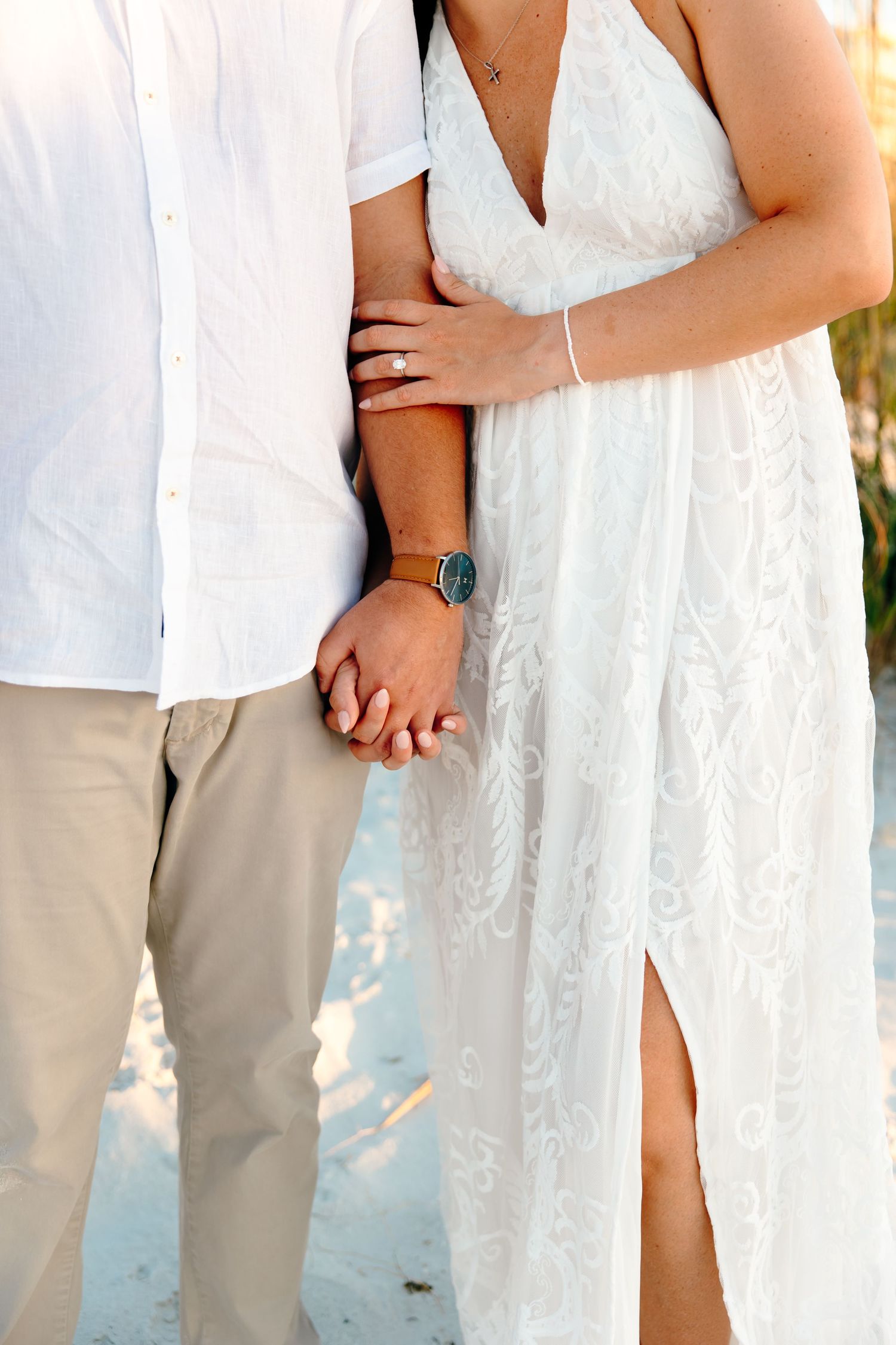Couple holding hands on beach, woman in white lace dress with engagement ring, man in white shirt and khaki pants.