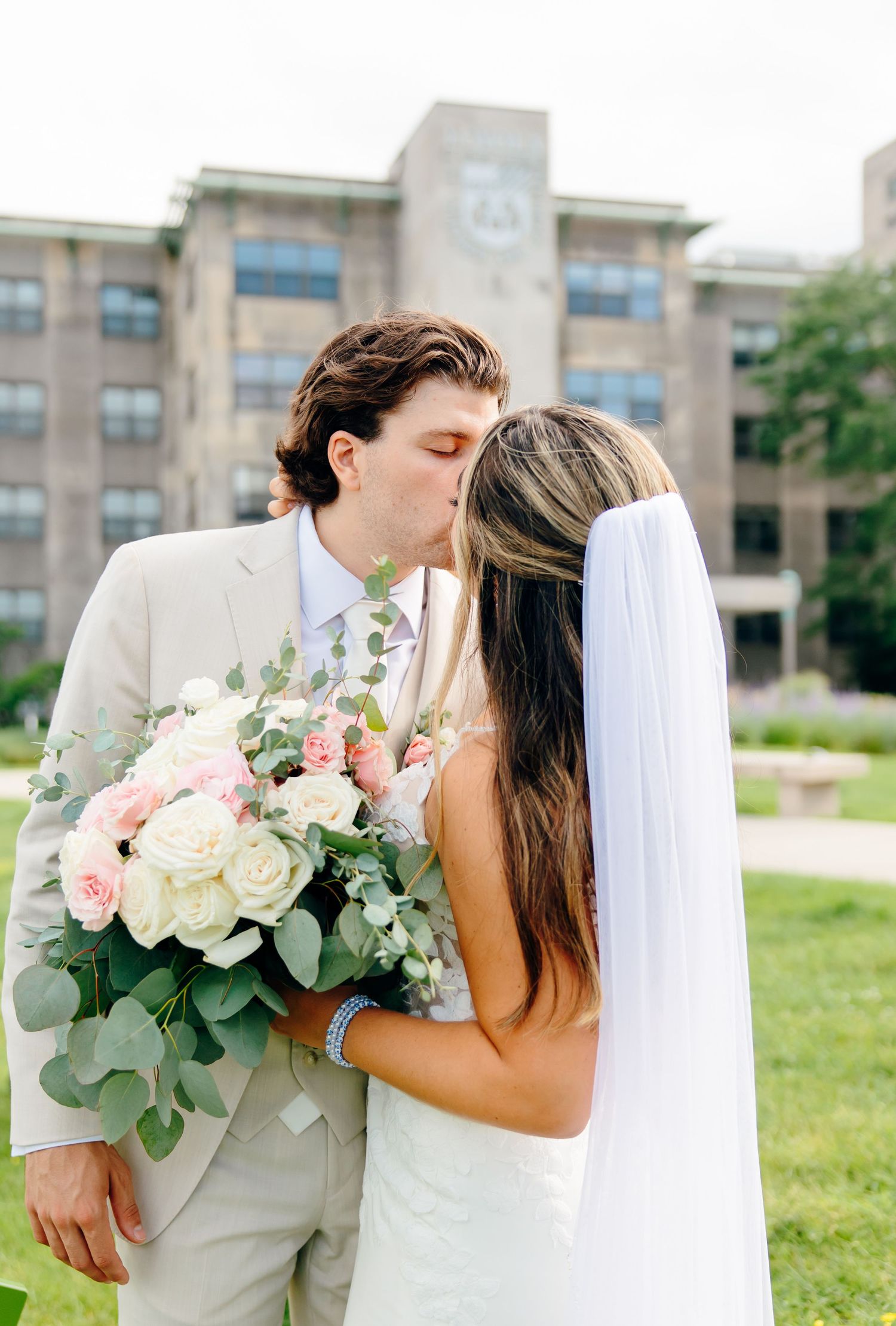 Bride and groom sharing a kiss outdoors, bride holding a bouquet of white and pink roses with greenery, wearing a veil.