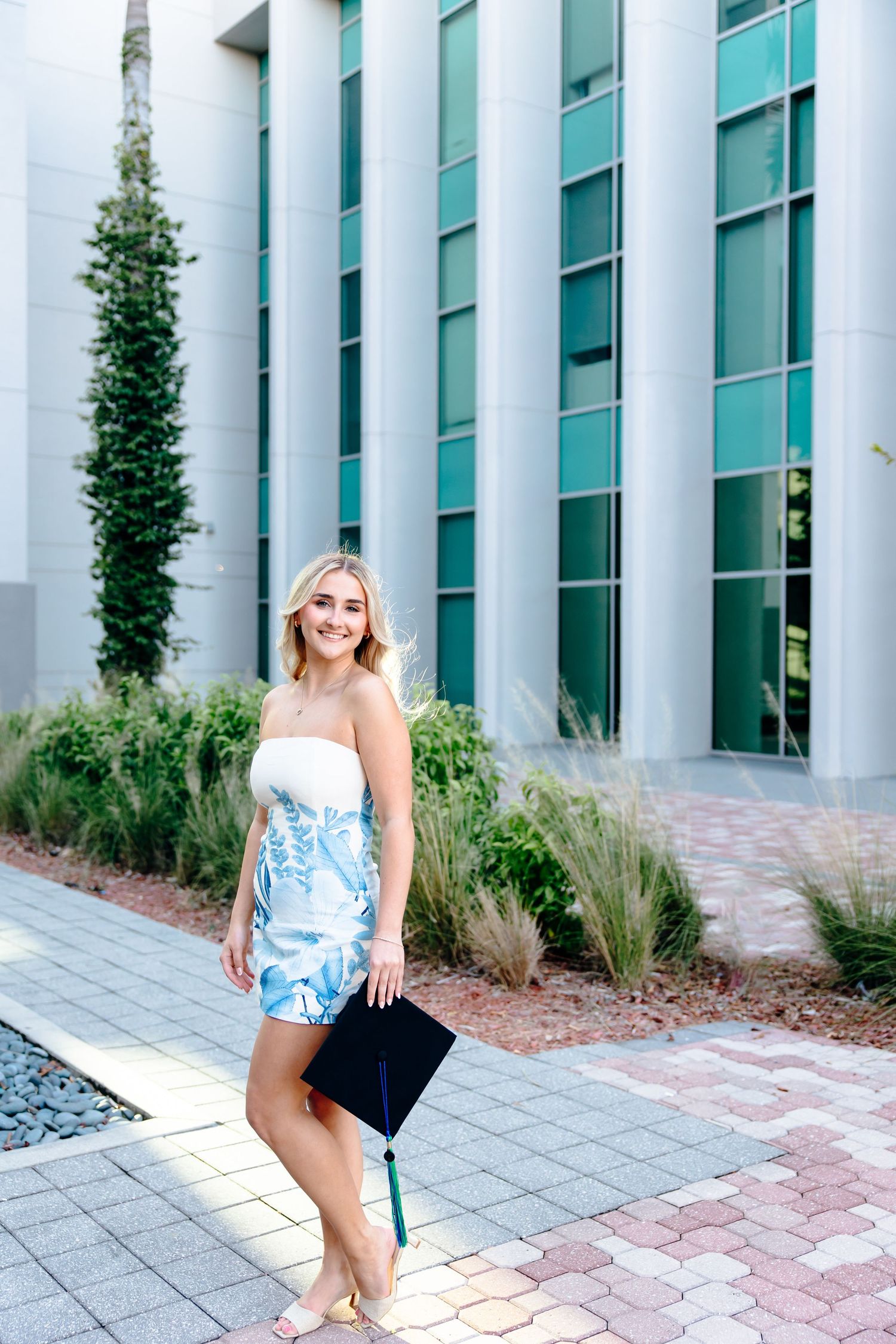 Smiling blonde graduate in blue floral dress holding cap outside modern university building on brick pathway.