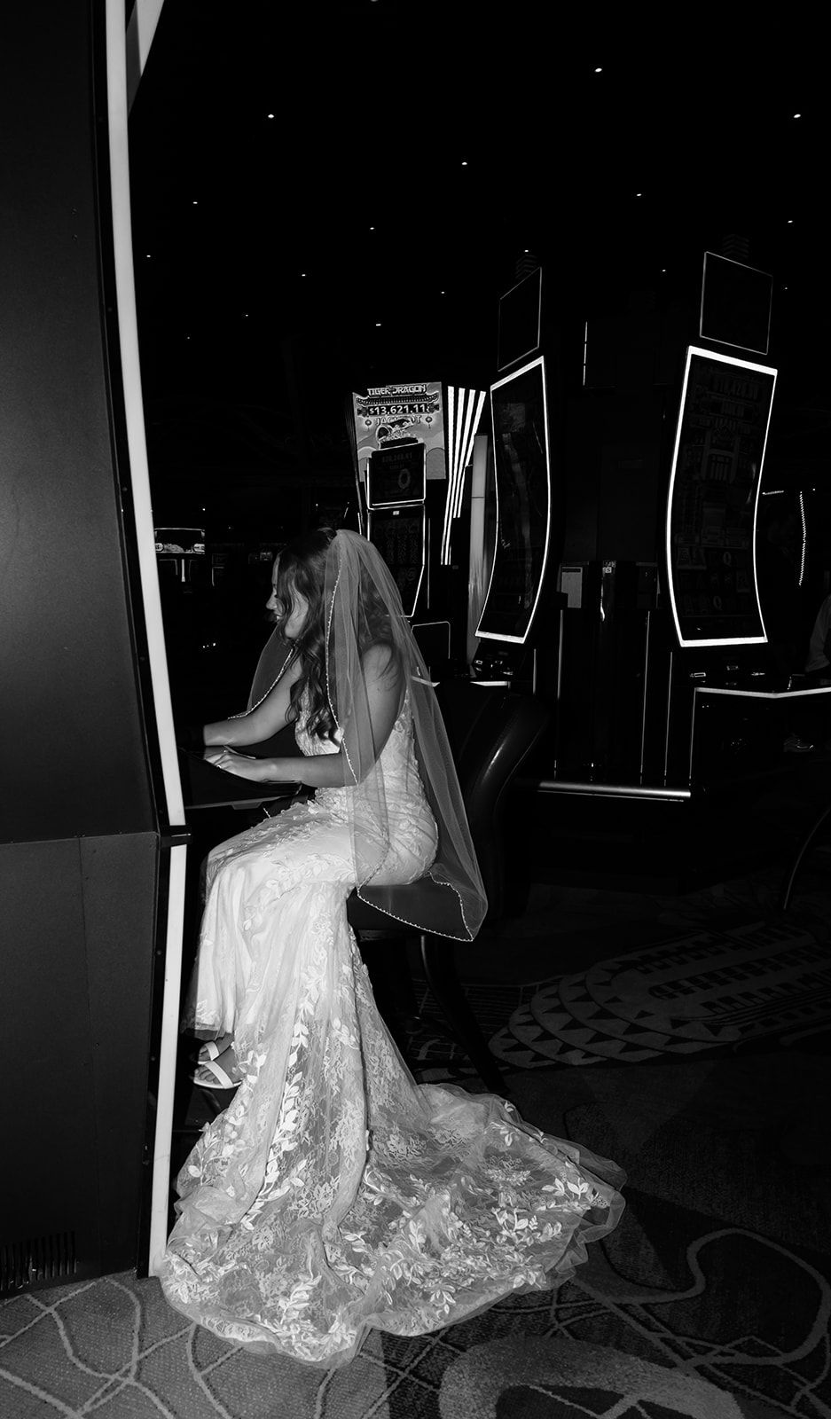 Black and white photo of a bride in lace wedding dress and veil sitting alone in a dimly lit casino.