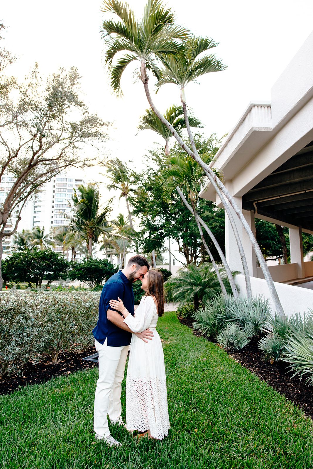 Couple sharing a romantic moment in a lush tropical garden with palm trees and a modern building backdrop.