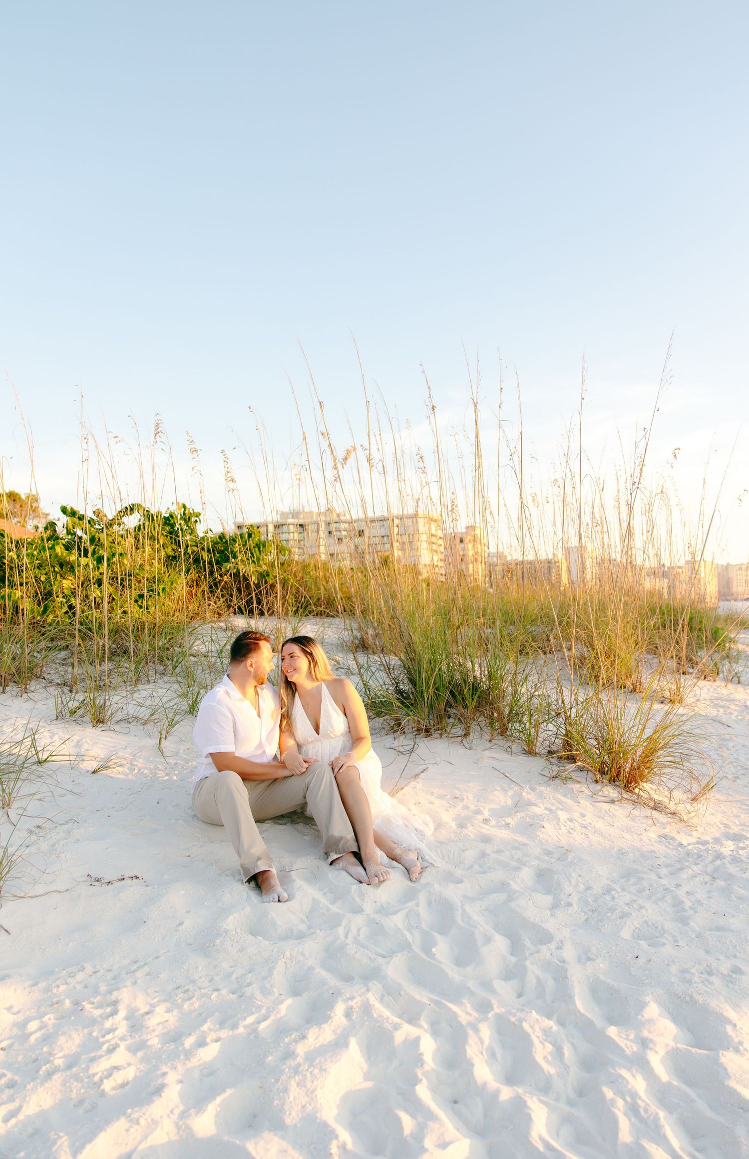 A couple sits together on white sandy beach dunes with tall sea grass and a sunset glow in the background.
