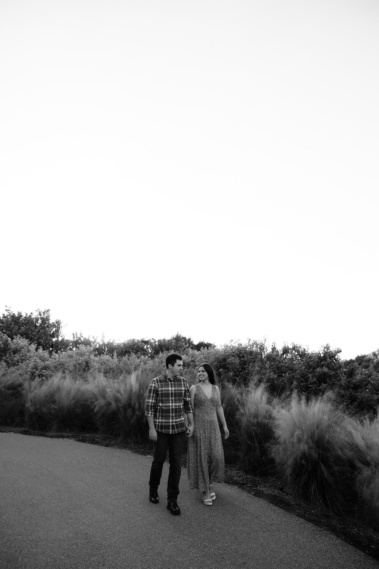 Black and white photo of a couple walking along a path surrounded by tall grasses at dusk.