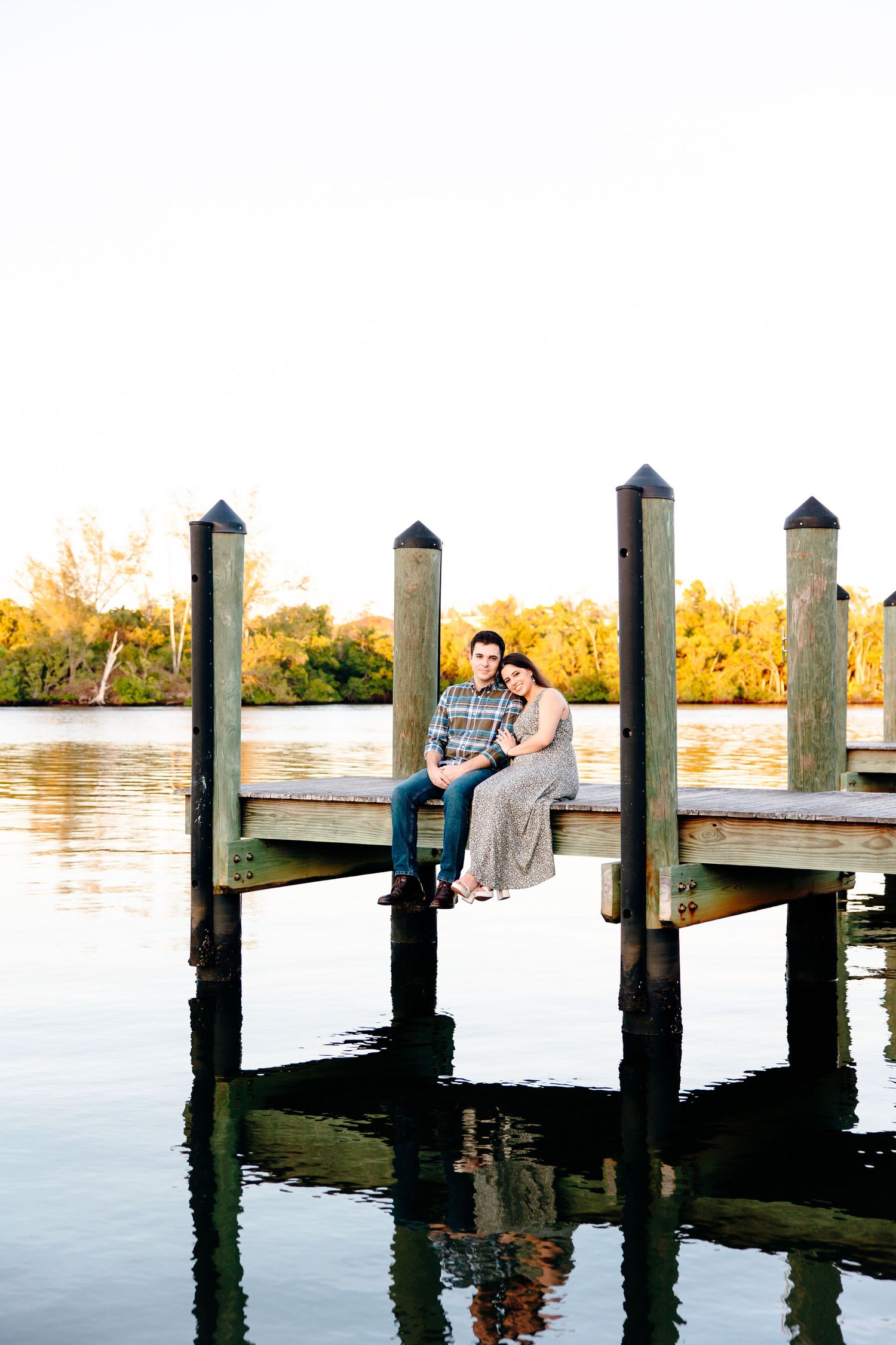 Couple sitting closely together on a wooden dock over calm water, surrounded by lush trees at golden hour.