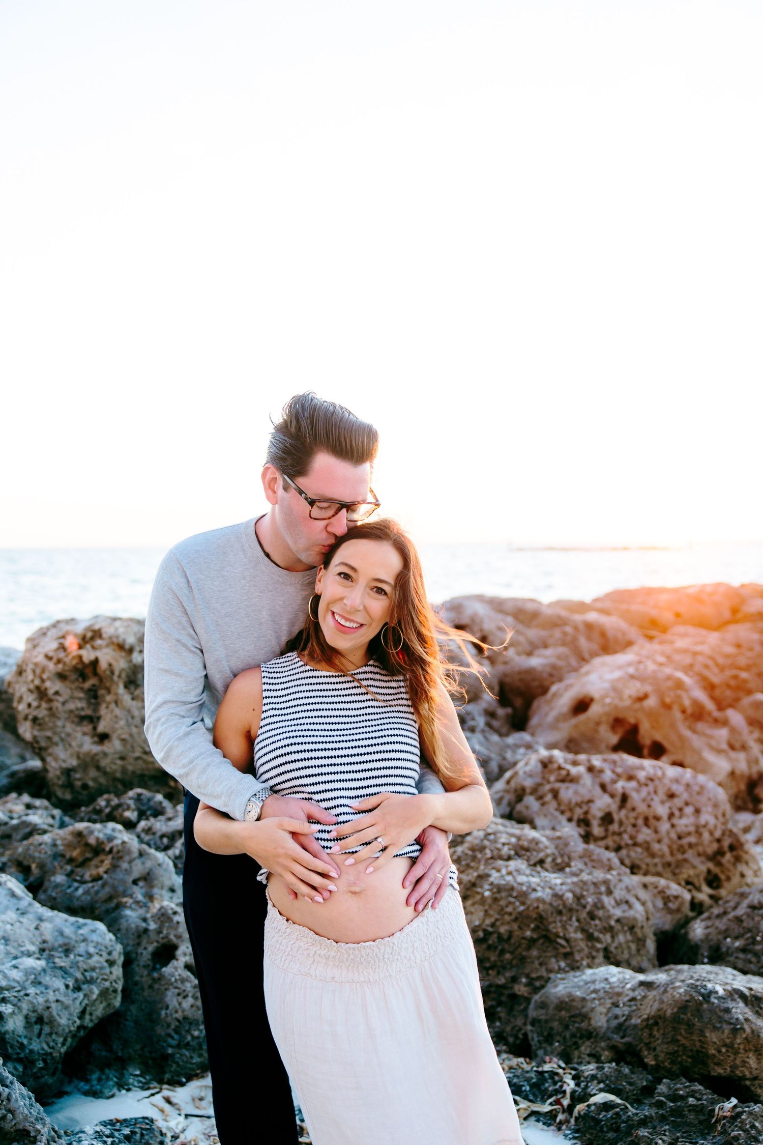 A couple poses on rocky beach at sunset, man embracing pregnant woman from behind, both smiling warmly.