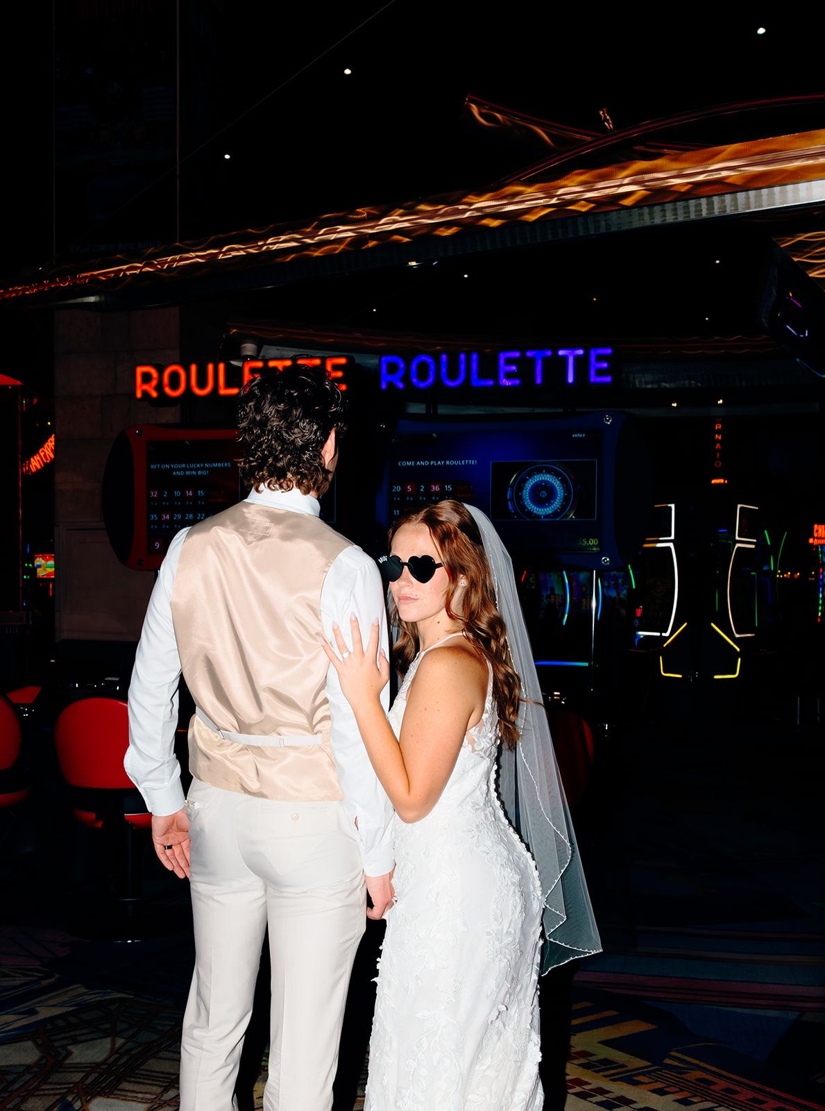 Bride and groom in white wedding attire posing together in front of neon roulette signs at a casino.