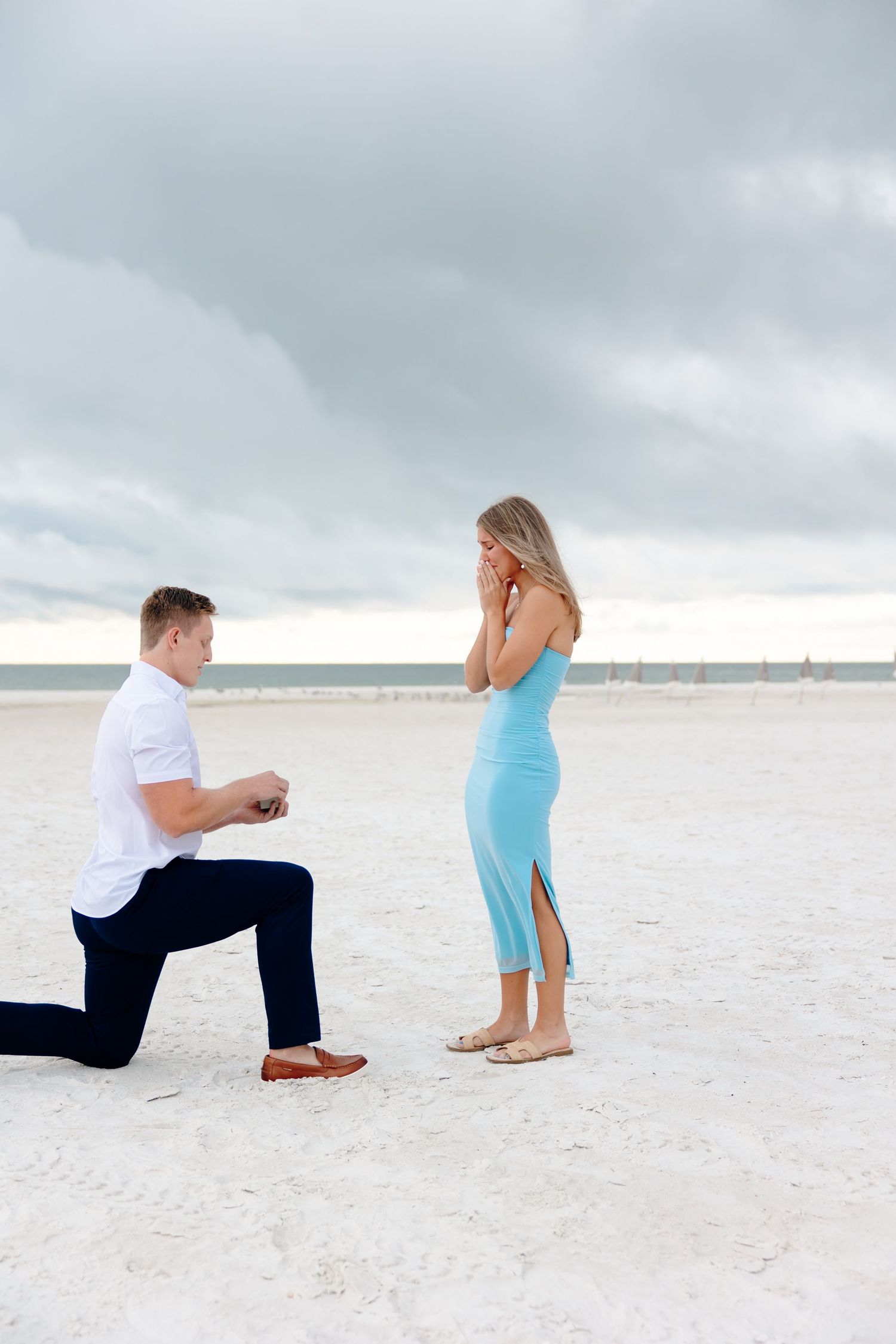 Man on one knee proposing to woman in blue dress on beach, she covers her mouth in surprise.