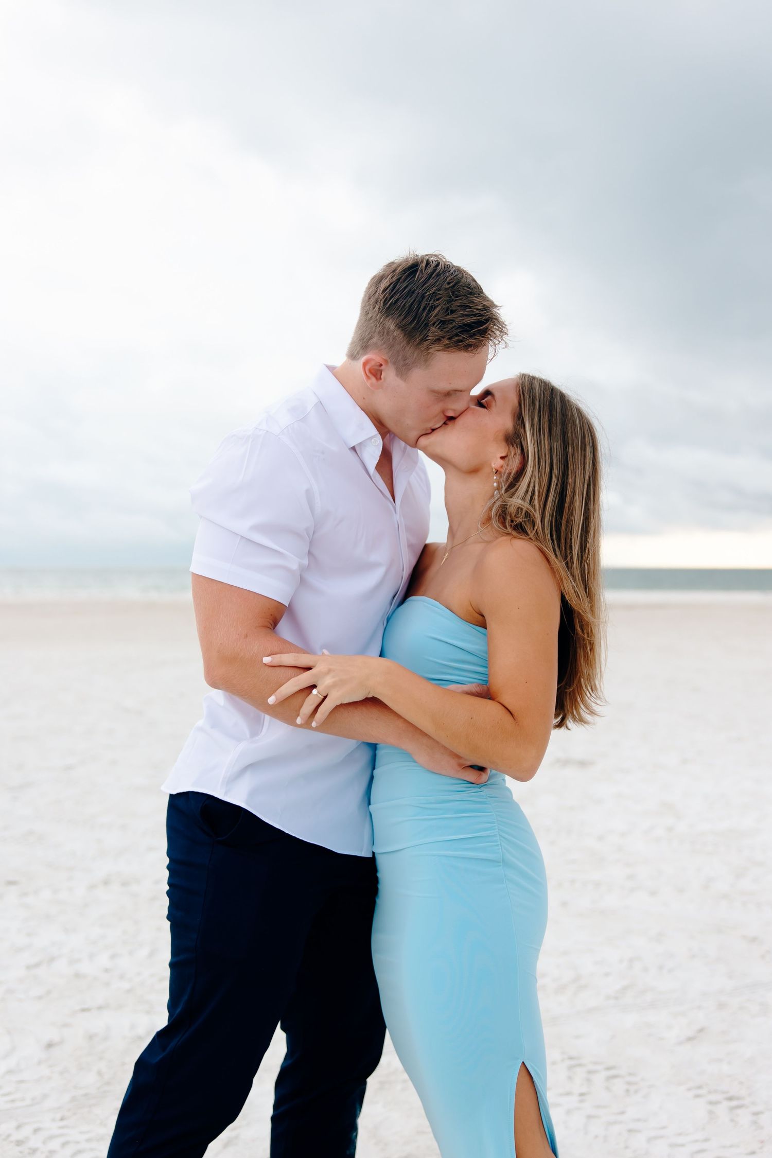 A couple in white and turquoise outfits share an intimate moment on a sandy beach under cloudy skies.
