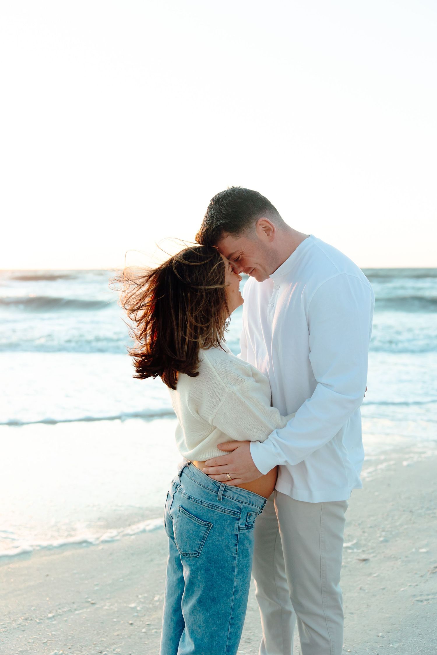 A romantic couple embraces on a windy beach, both wearing white tops and jeans, with ocean waves in the background.