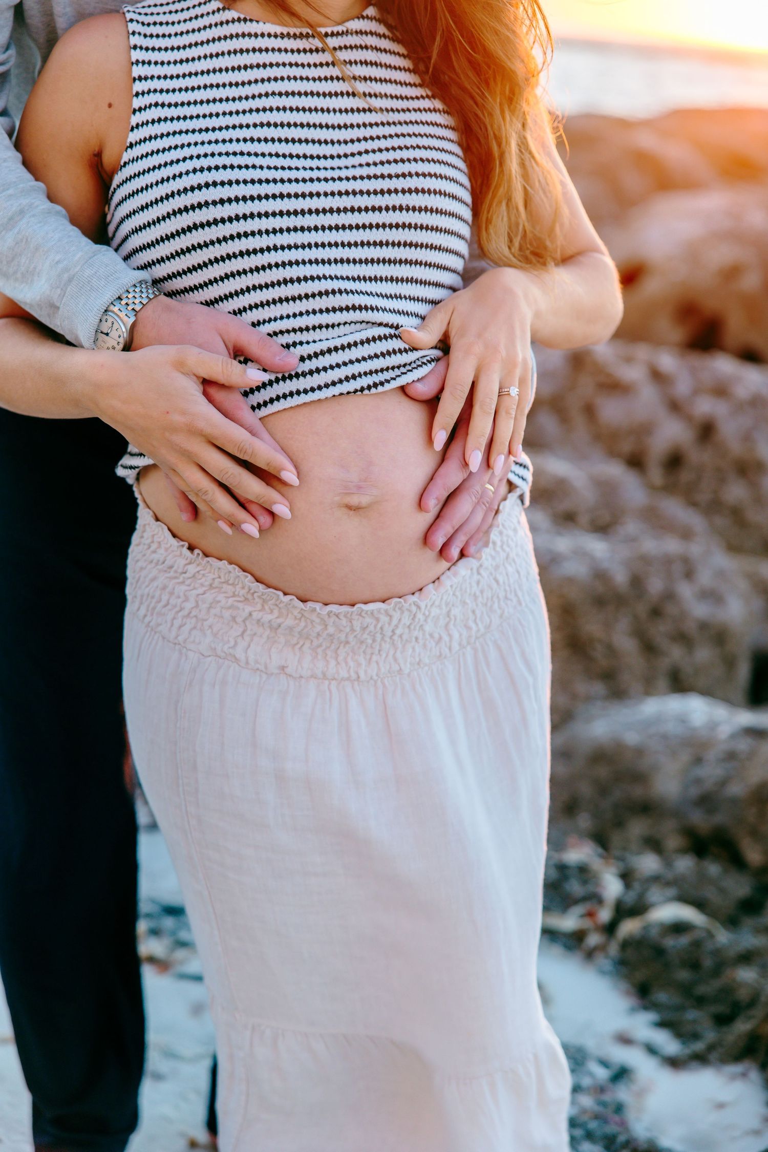 Couple cradling pregnant belly outdoors near rocky beach at sunset, woman in striped top and white skirt.