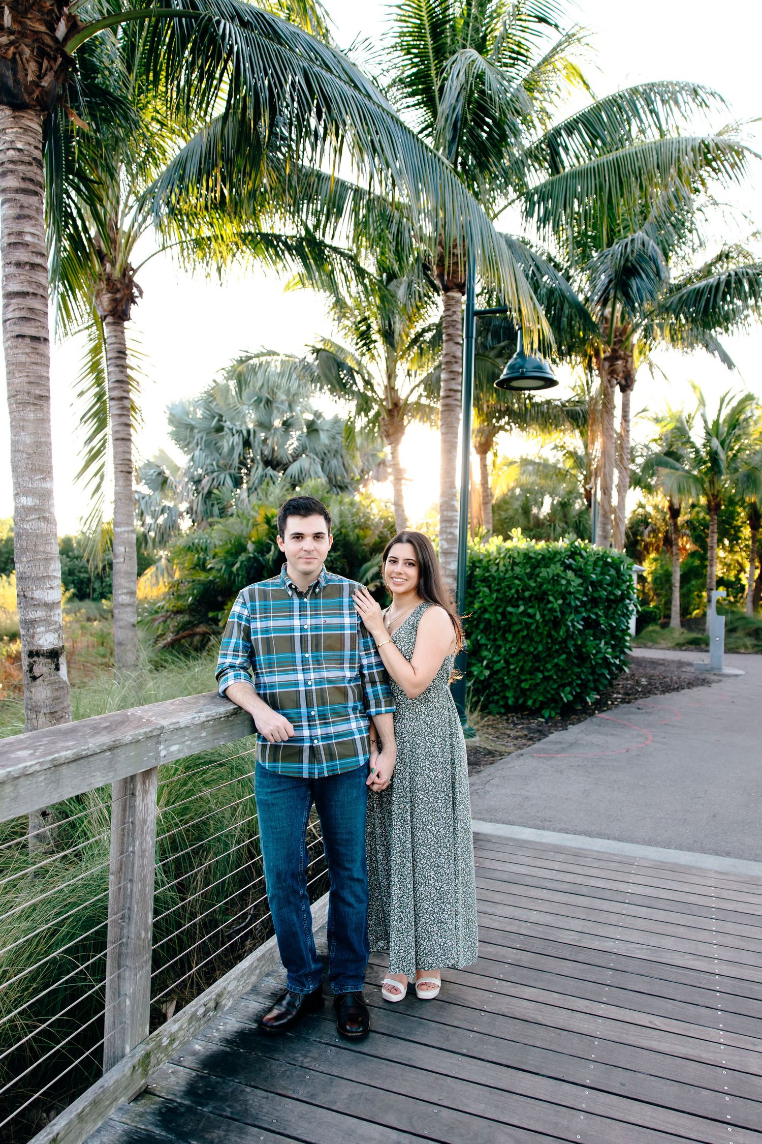 Couple posing on a wooden bridge surrounded by lush palm trees in a tropical park setting.