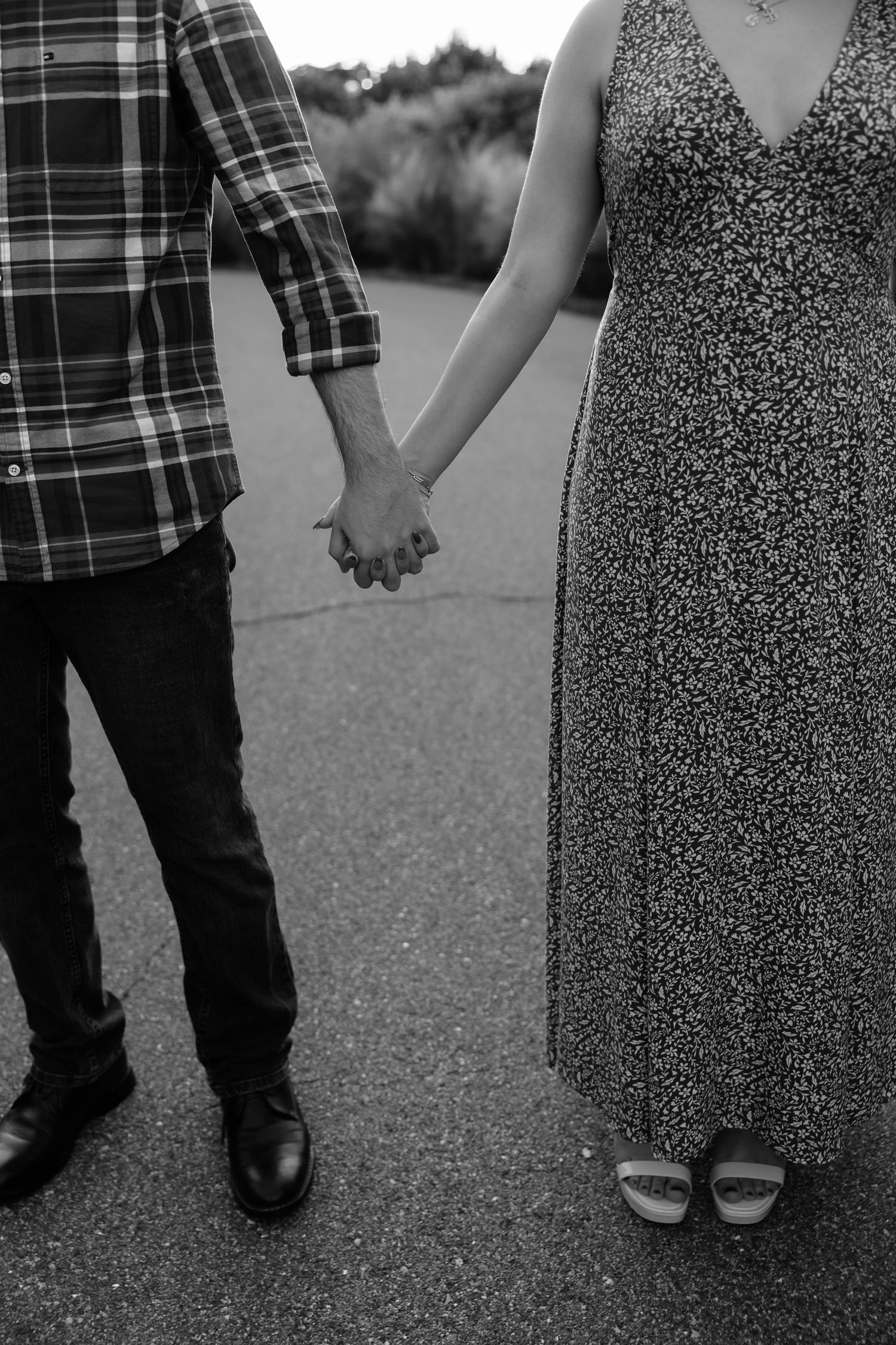 Black and white photo of a couple holding hands, man in plaid shirt and jeans, woman in floral dress, standing on pavement.