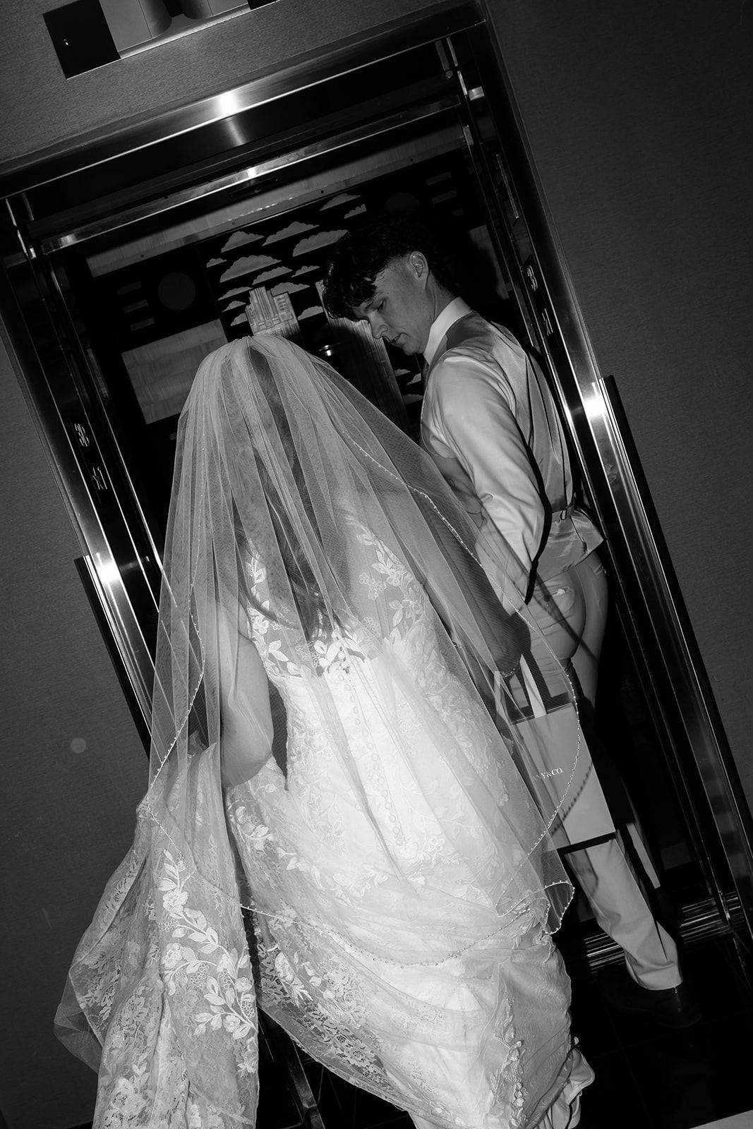 Black and white photo of bride in lace gown and veil kissing groom in elevator at wedding.