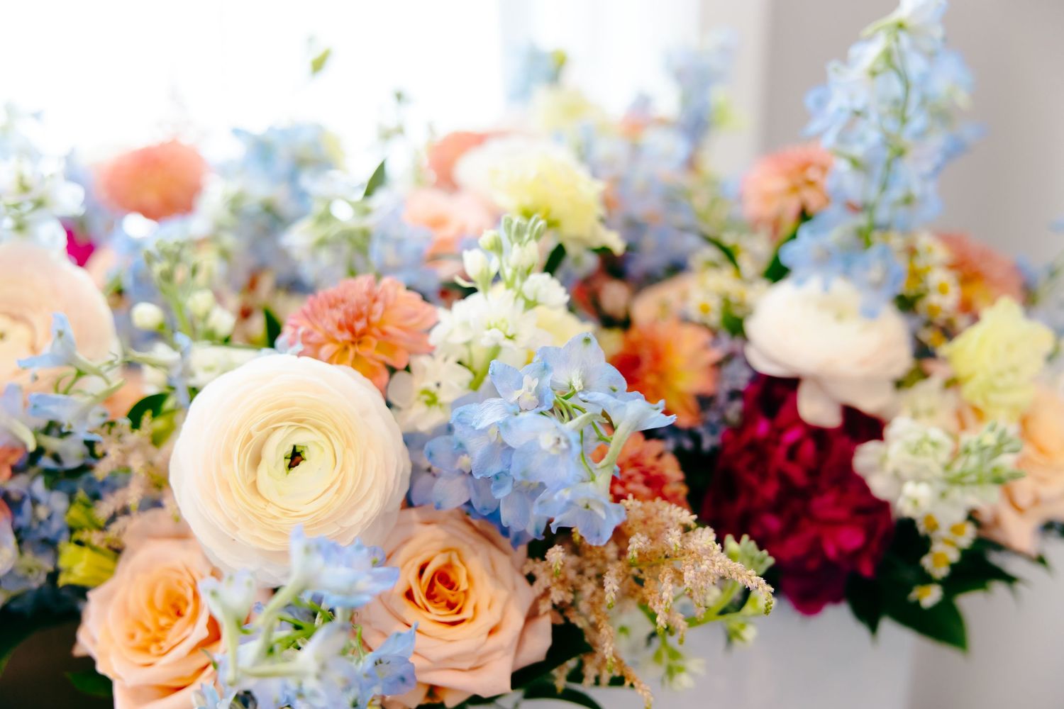 Colorful wedding bouquet featuring peach roses, white ranunculus, blue hydrangeas, and mixed blooms in soft pastel tones.