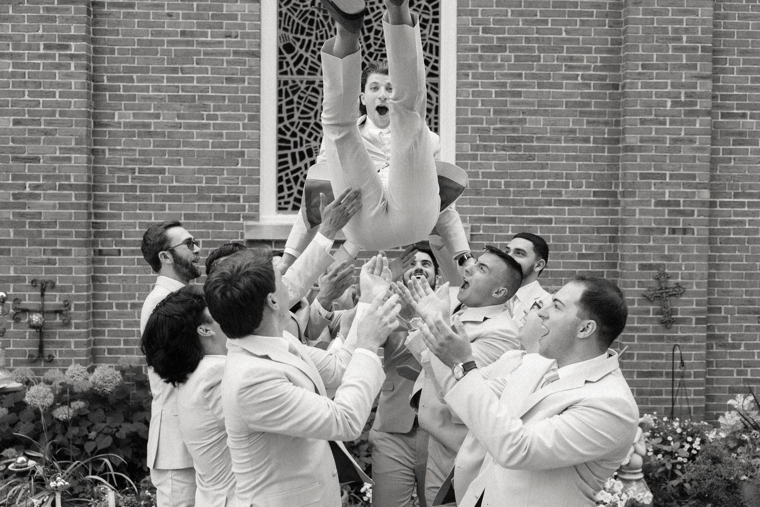 Groomsmen in white suits tossing a laughing groom into the air outside a brick church in a black and white photo.