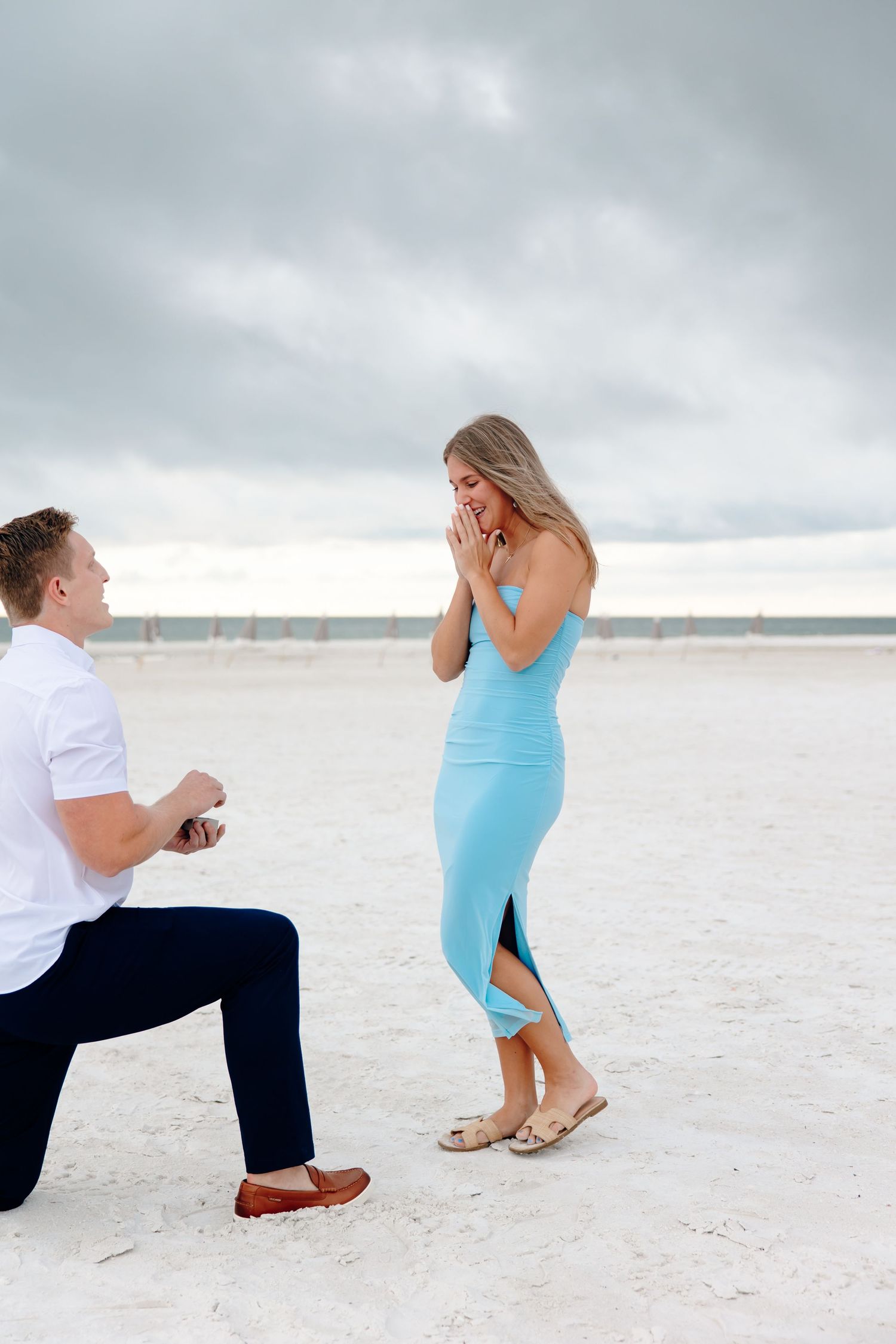 Couple during beach marriage proposal, man kneeling with ring as woman reacts with hands over mouth.