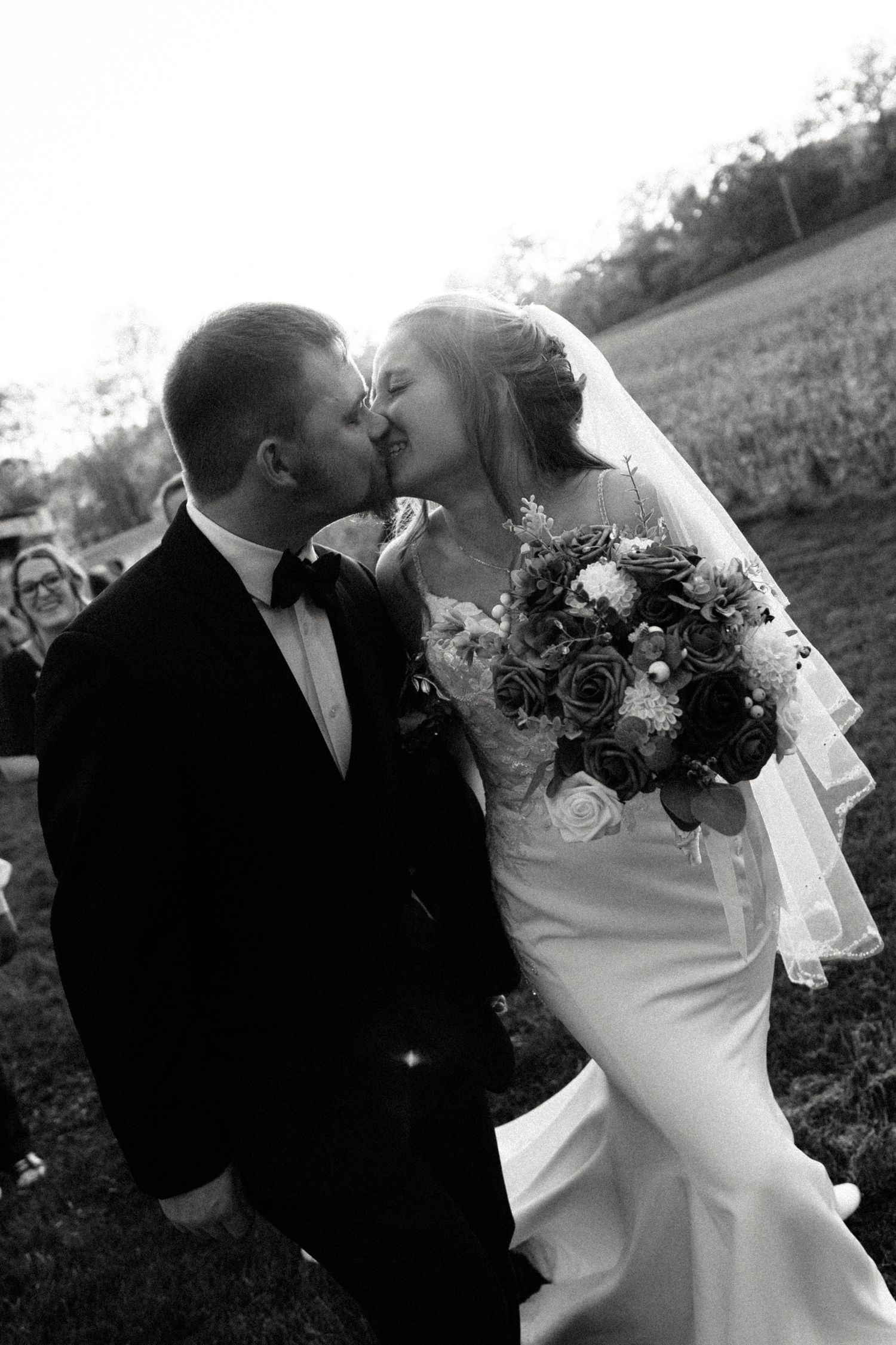 Black and white photo of a bride and groom sharing a kiss outdoors, bride holding a bouquet of roses in a wedding gown.