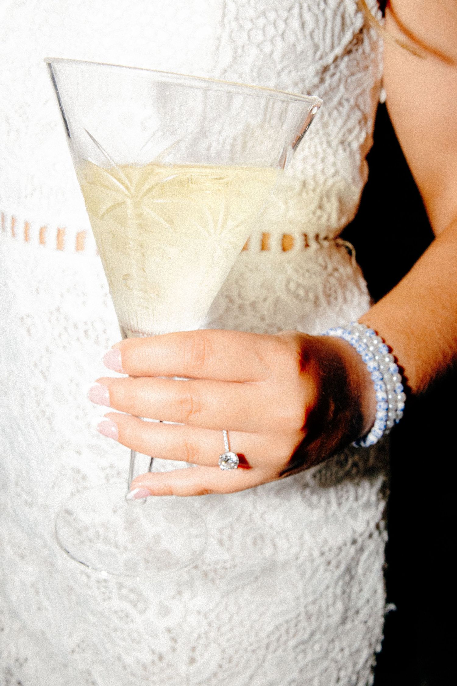 Bride in white lace dress holding a martini glass with diamond ring and pearl bracelet.