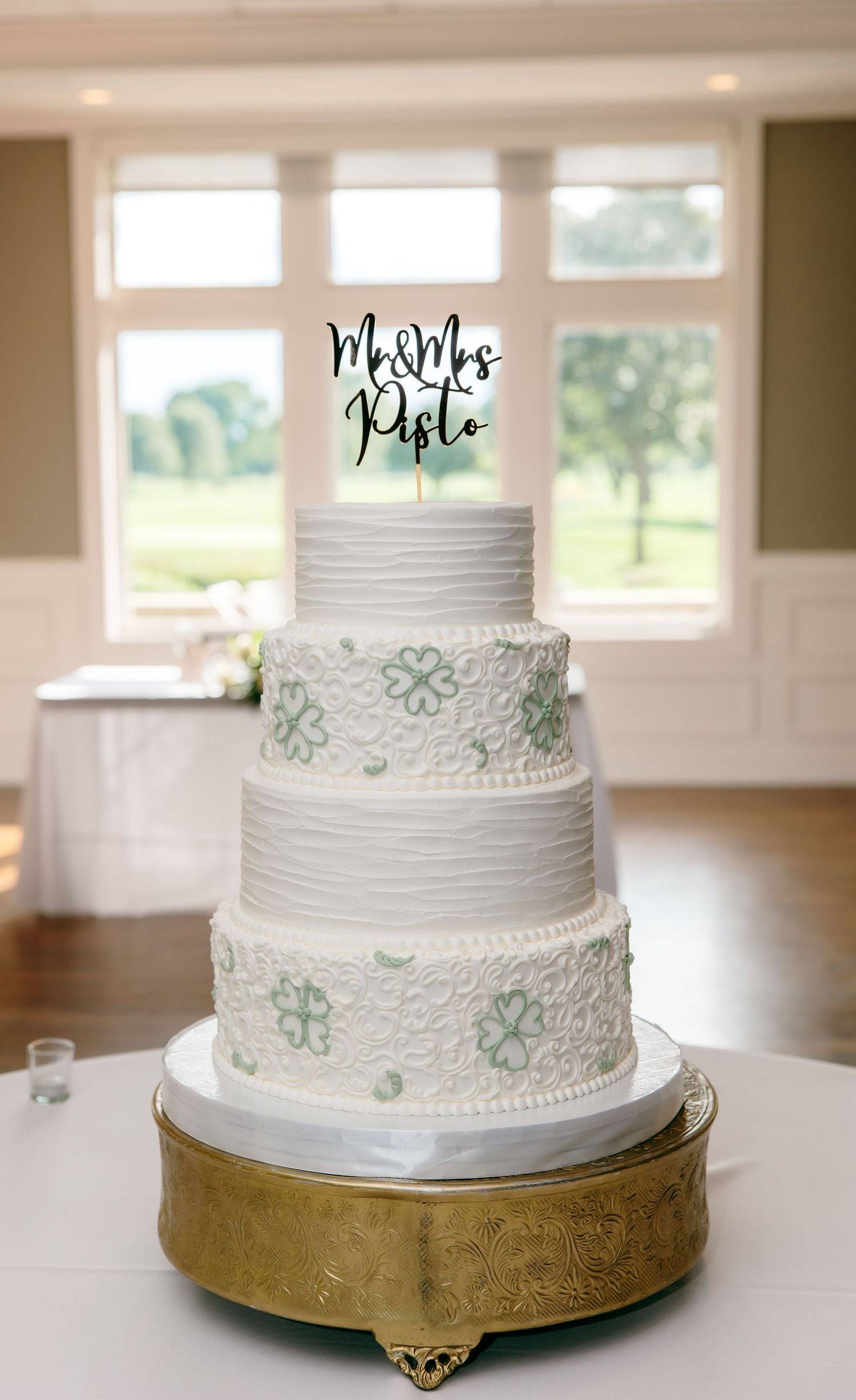 Elegant white tiered wedding cake with mint green floral accents displayed on a gold stand in a bright room.