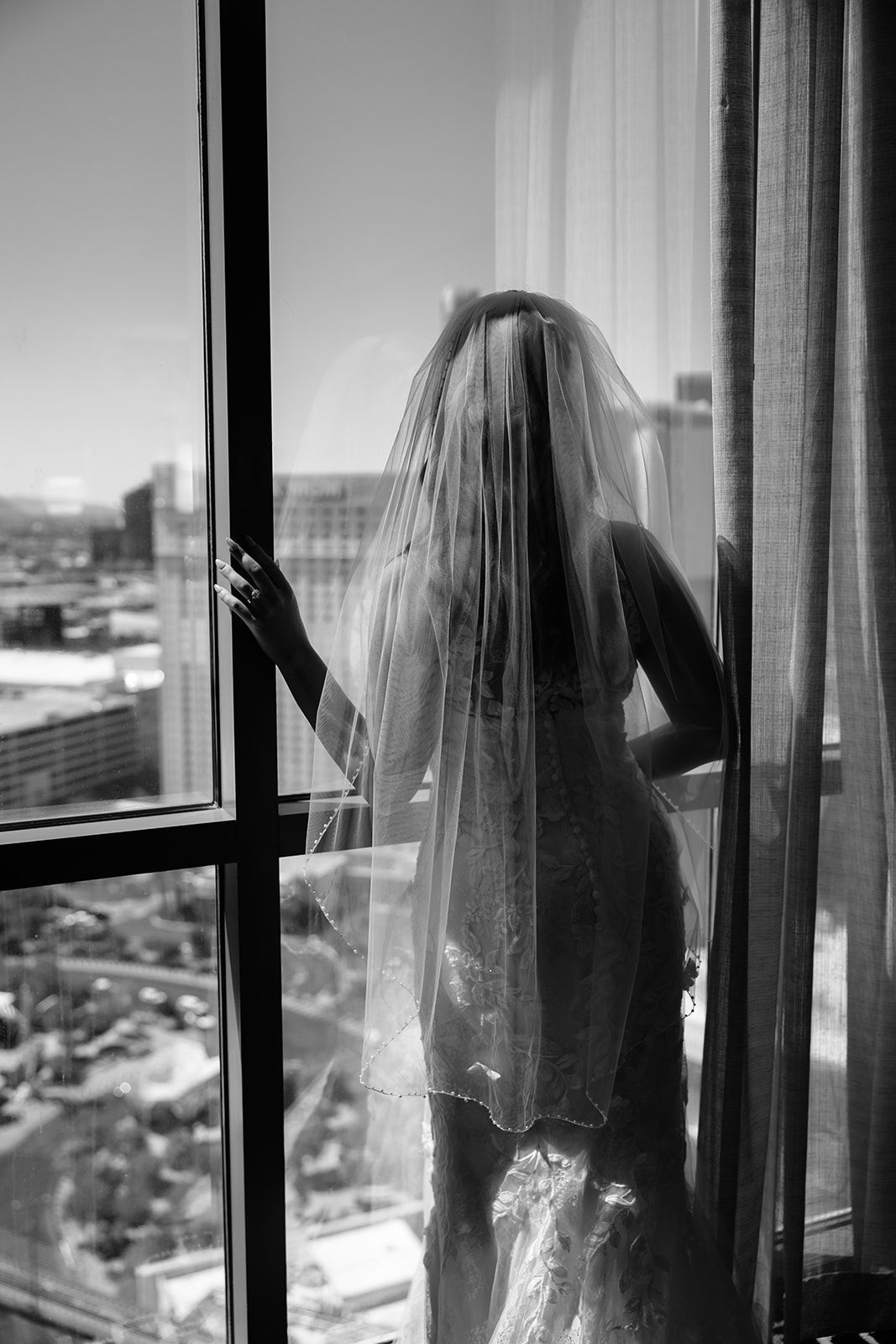Bride in wedding dress and veil stands at high-rise window overlooking city skyline, black and white photo.