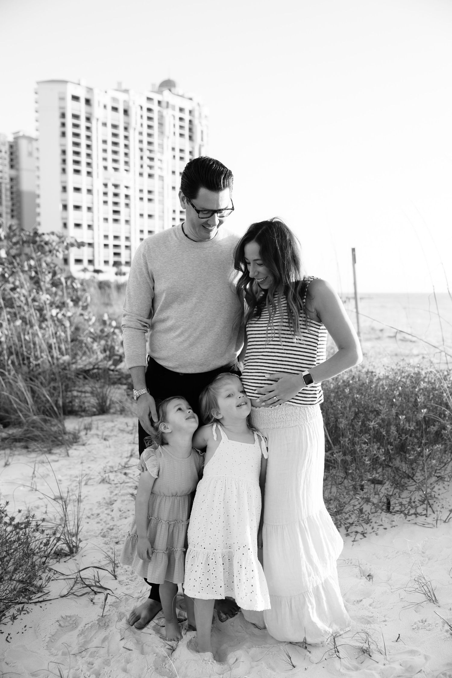 Black and white family portrait on beach with father, pregnant mother, and two young daughters hugging.