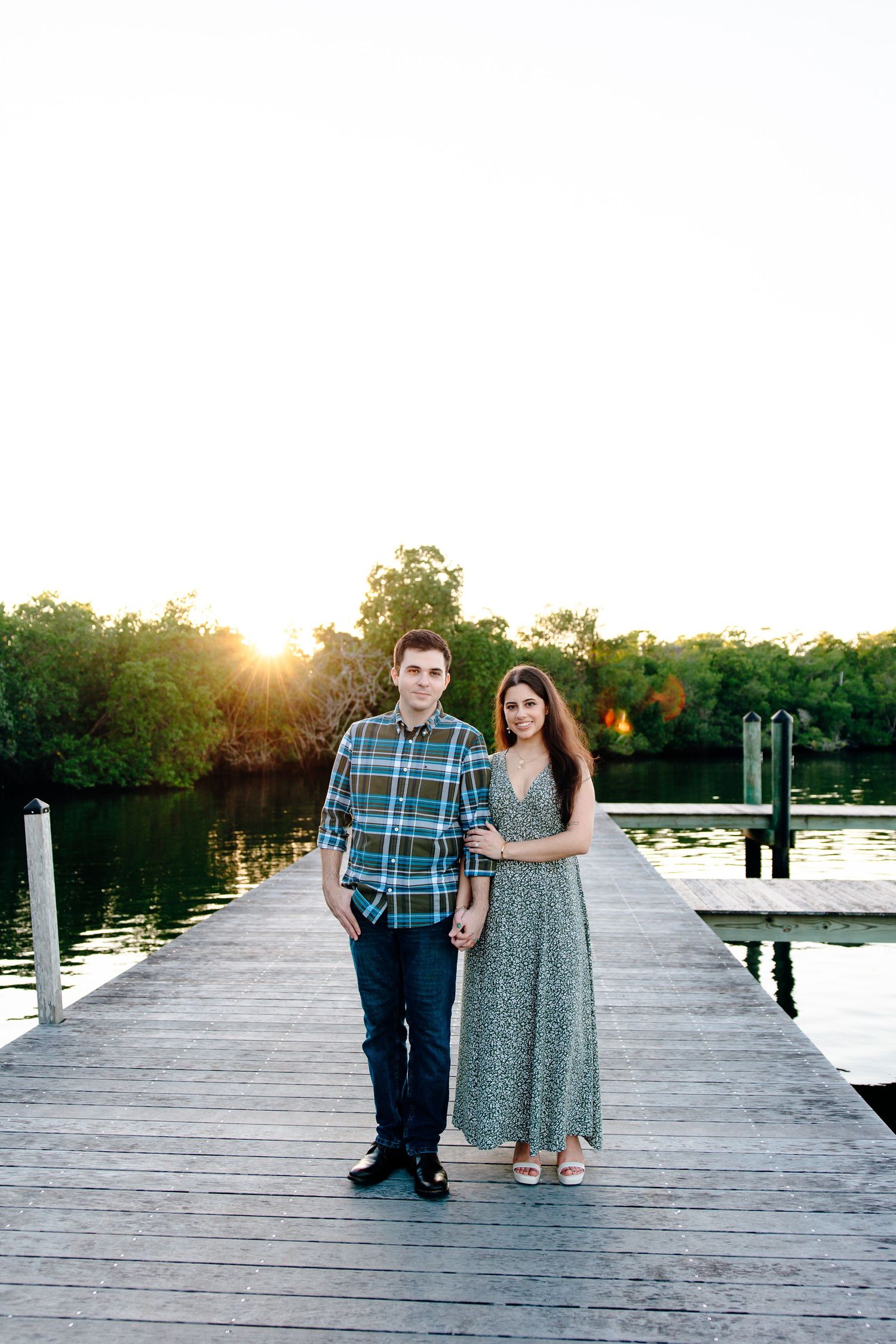Couple standing together on a wooden dock at sunset, surrounded by lush greenery and calm water.