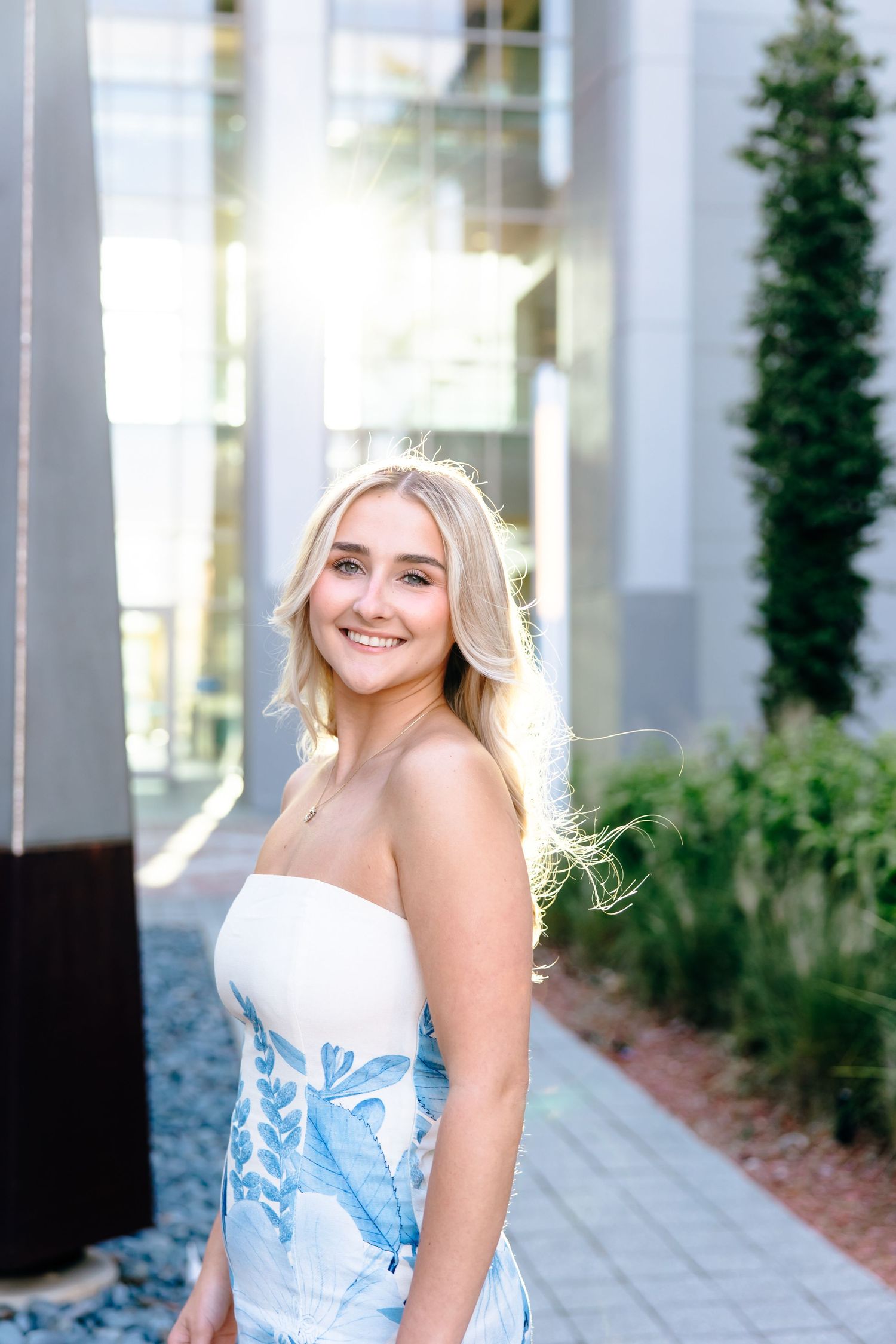 Smiling blonde woman in a strapless blue floral dress posing outdoors near modern building with greenery.