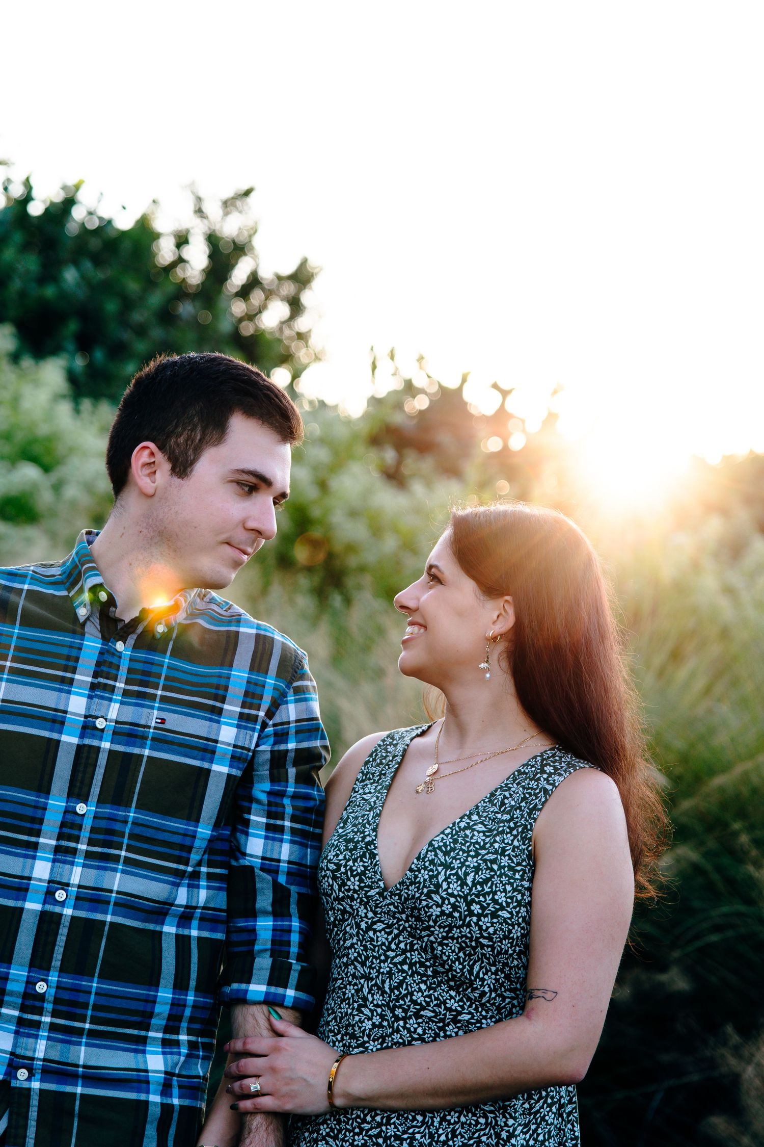 Couple sharing a romantic moment outdoors at golden hour, man in plaid shirt and woman in floral dress smiling at each other.