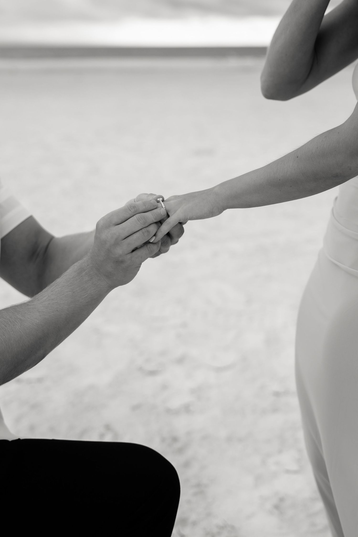 Black and white photo of a man on one knee placing an engagement ring on a woman's finger on a beach.